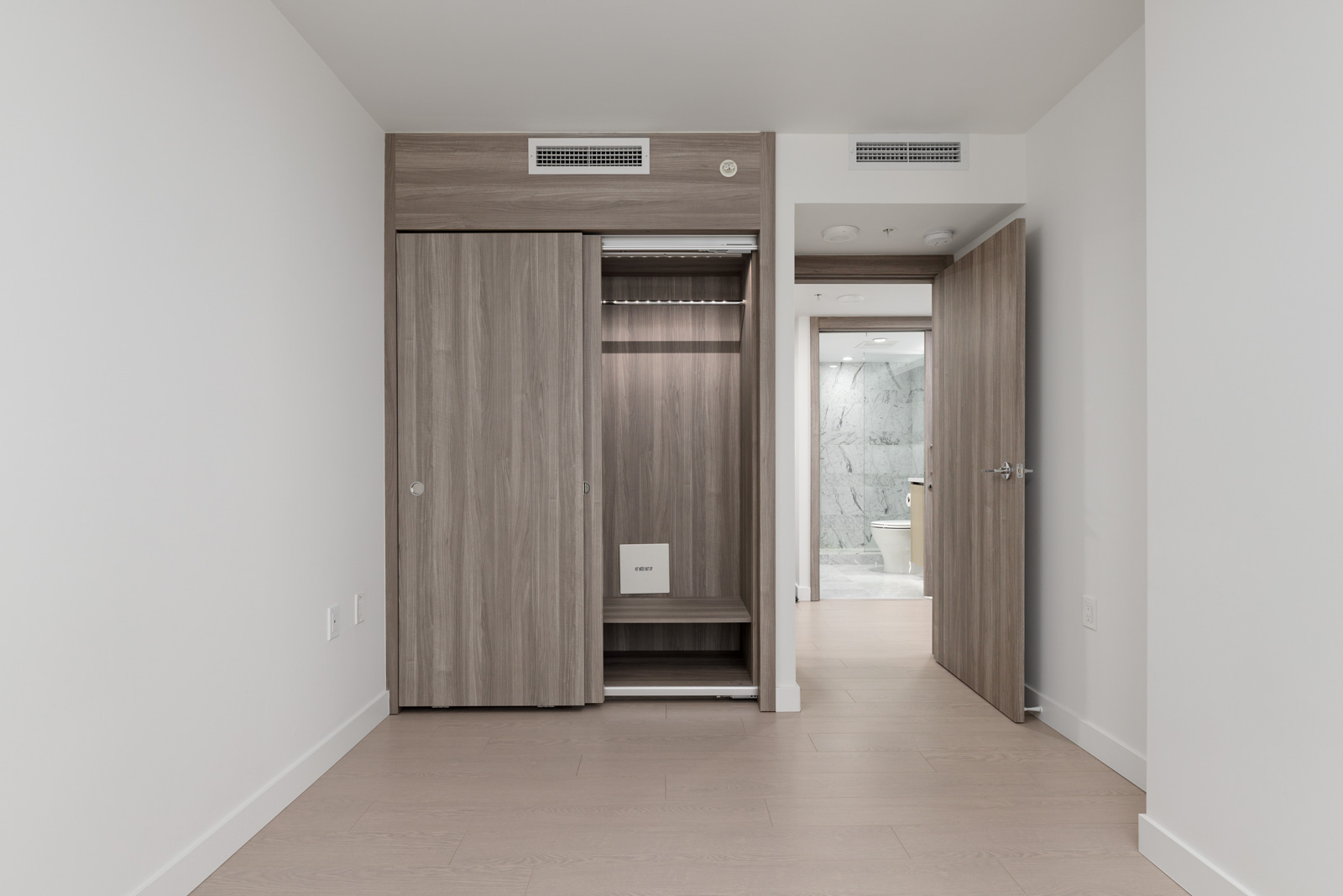Empty modern bedroom with light wood flooring, an open closet, and an open door leading to a bathroom with white marble walls.