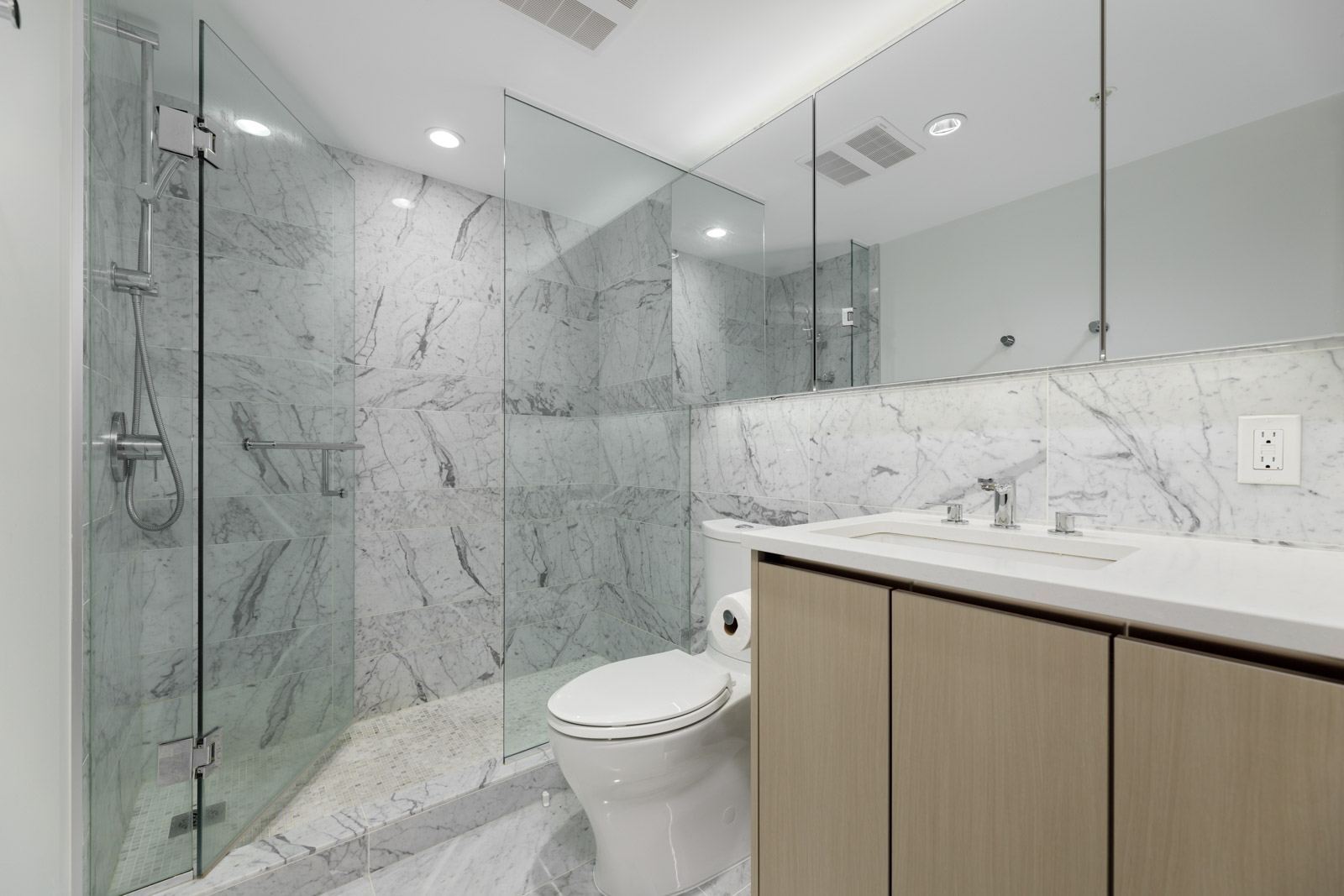Modern bathroom with marble walls and floors, glass-enclosed shower, toilet, double sink vanity with wood cabinets, and recessed lighting.