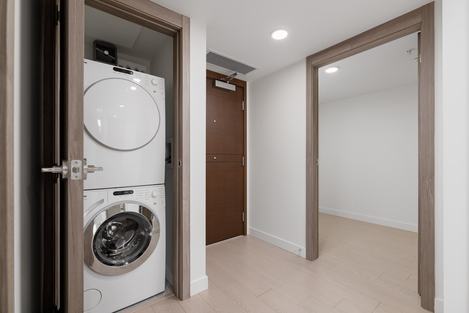 A stacked washer and dryer set is installed in a closet next to a hallway with light wood floors and open doors leading to other rooms.