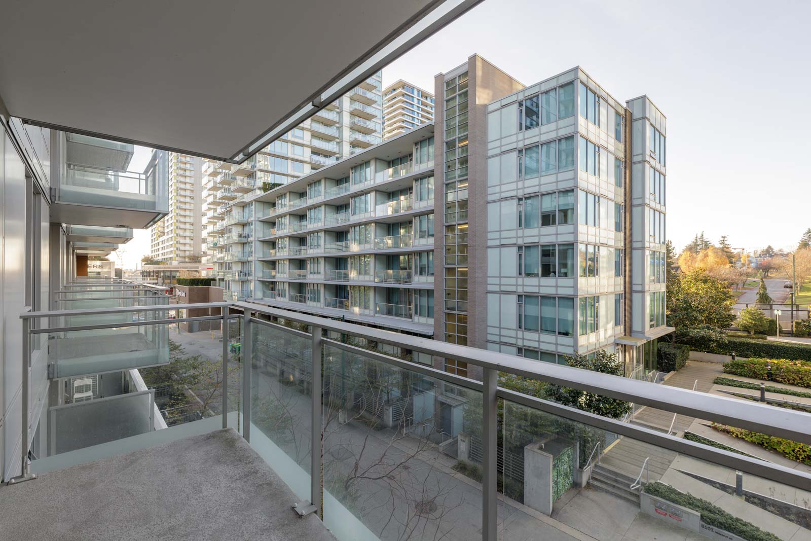 View from a modern apartment balcony overlooking glass and concrete residential buildings, with greenery and a clear sky in the background.