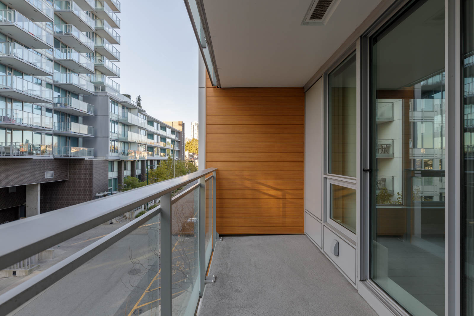 A modern apartment balcony with glass railing, concrete floor, and wood paneling, overlooking nearby buildings and a street below.