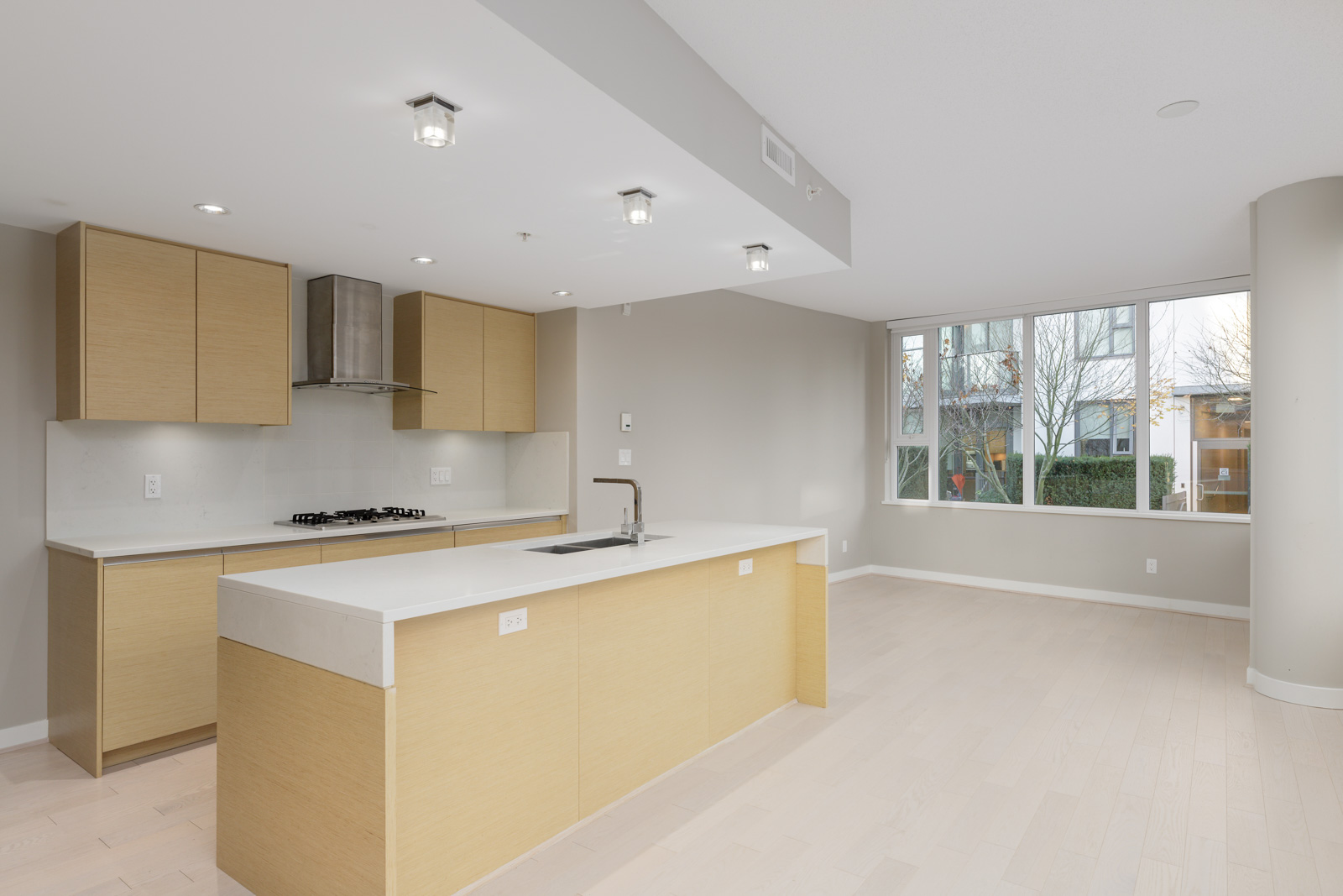Modern kitchen with light wood cabinets, white countertops, built-in gas stove, and an island; open layout connects to a bright living area with large windows.