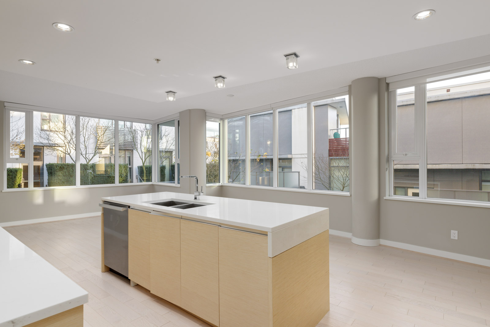 Modern kitchen with light wood cabinets, white countertops, a central island with a sink, and large windows providing ample natural light.