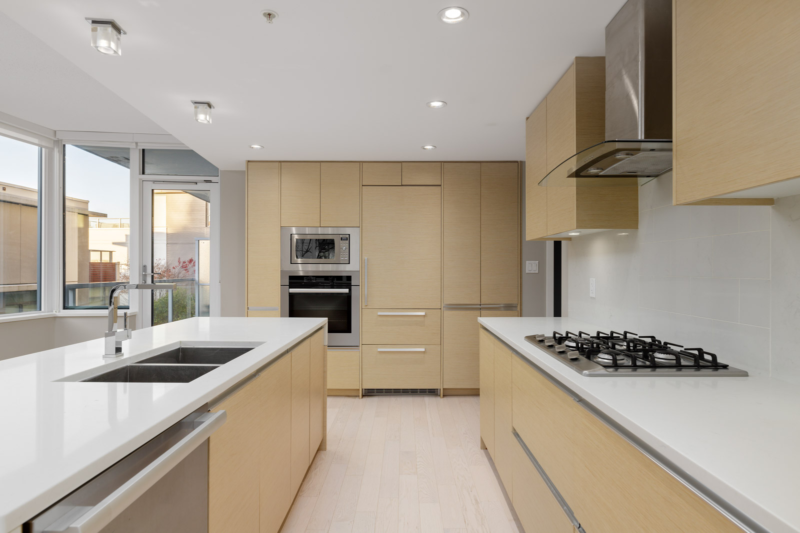 Modern kitchen with light wood cabinets, white countertops, built-in oven and microwave, gas cooktop, stainless steel hood, and large windows providing natural light.