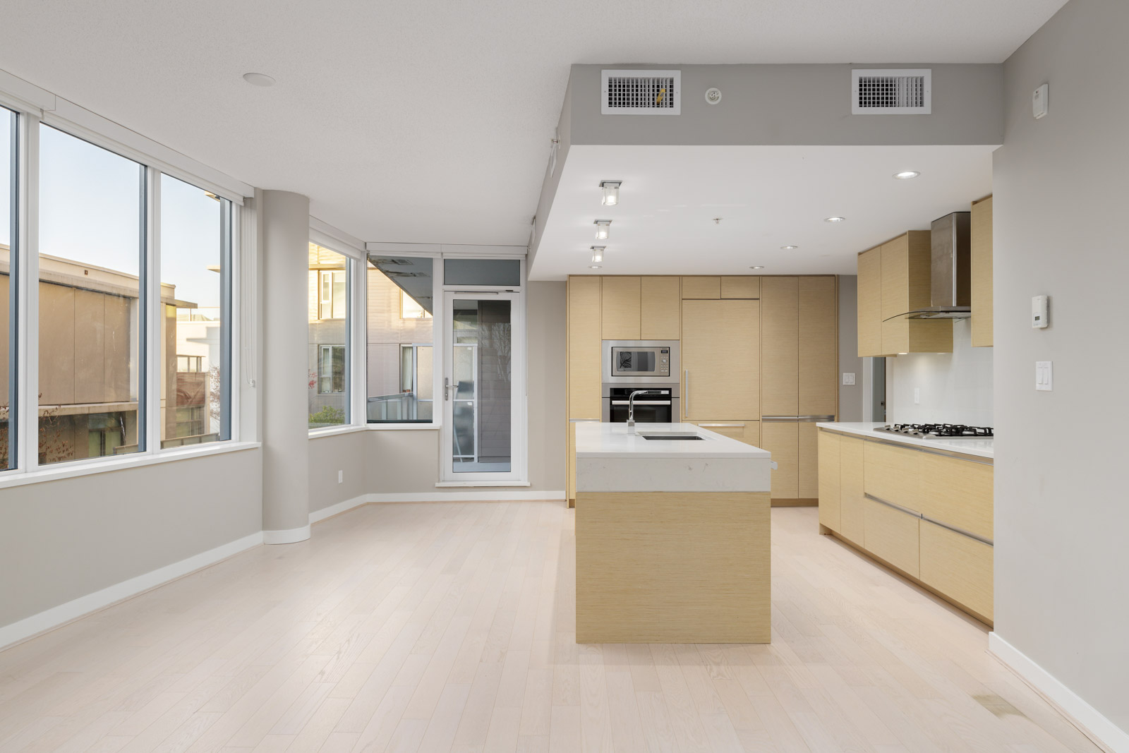 Modern, unfurnished kitchen with light wood cabinets, center island, built-in appliances, and large windows allowing natural light into an open living area.