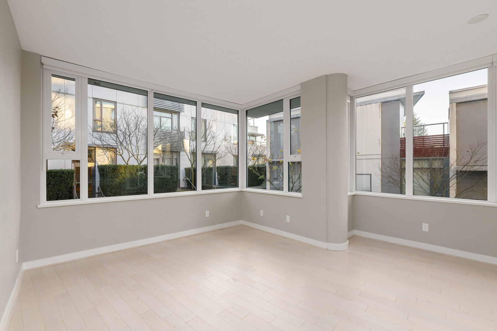Empty corner room with large windows, light-colored wood floors, neutral walls, and views of nearby buildings and trees outside.