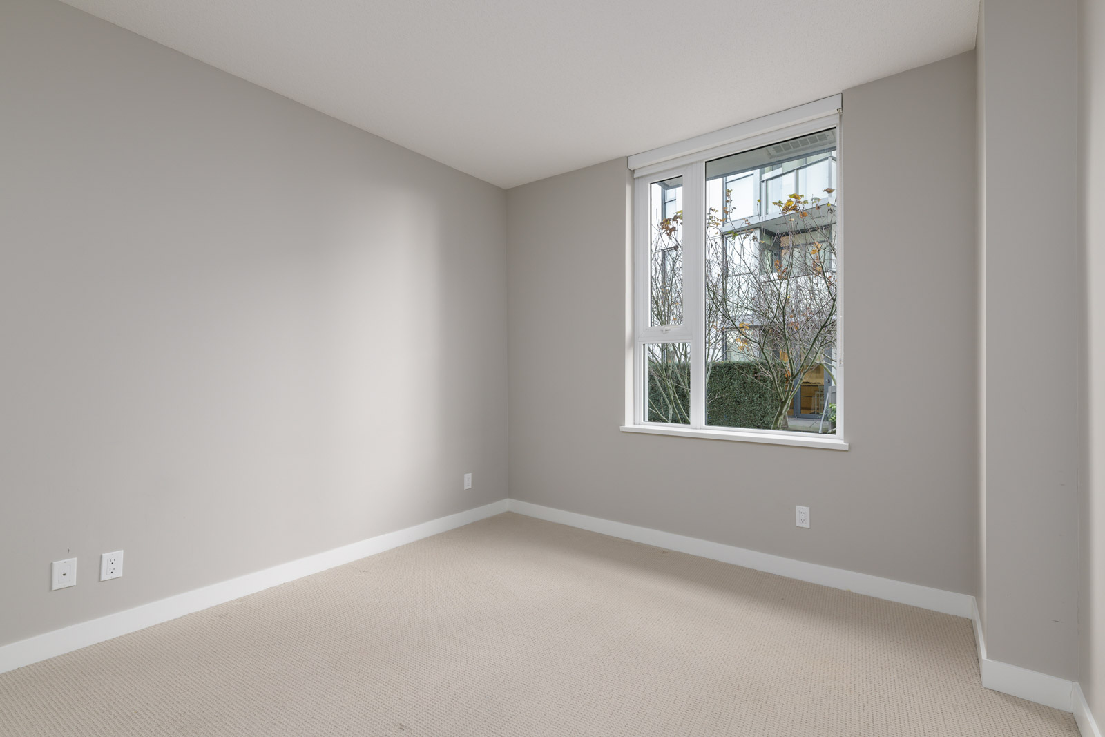Empty room with beige carpet, light gray walls, and a large window with a view of trees and a building exterior. There are power outlets on the walls.