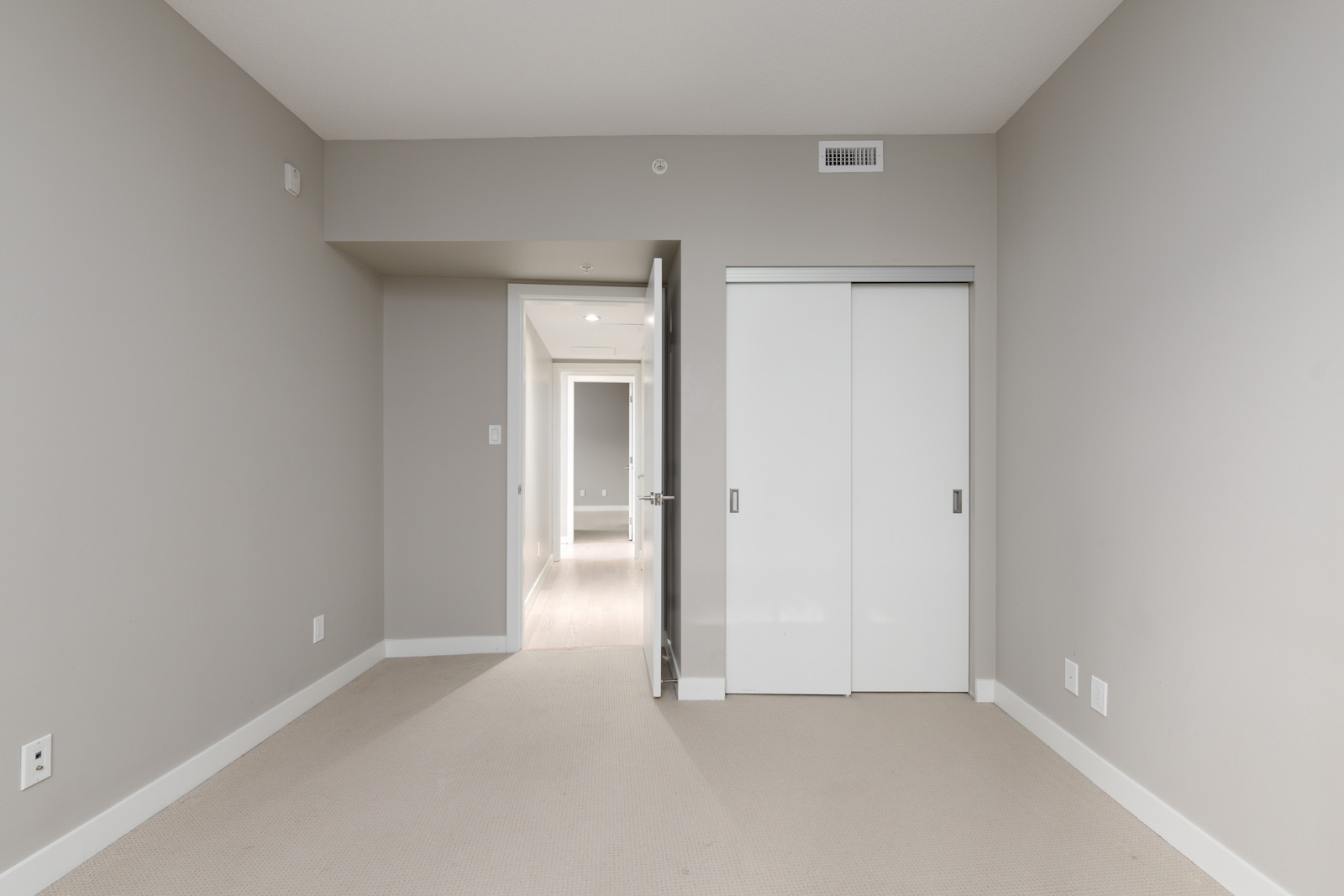 Empty, unfurnished room with beige carpet, light grey walls, white closet doors, and view of a hallway with doors and natural light in the distance.