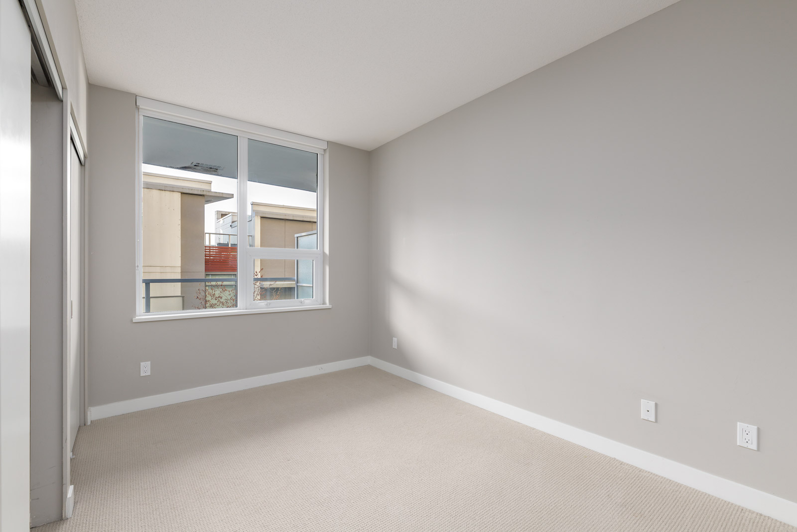 Empty room with beige carpet, light gray walls, a large window, and electrical outlets visible on one wall.