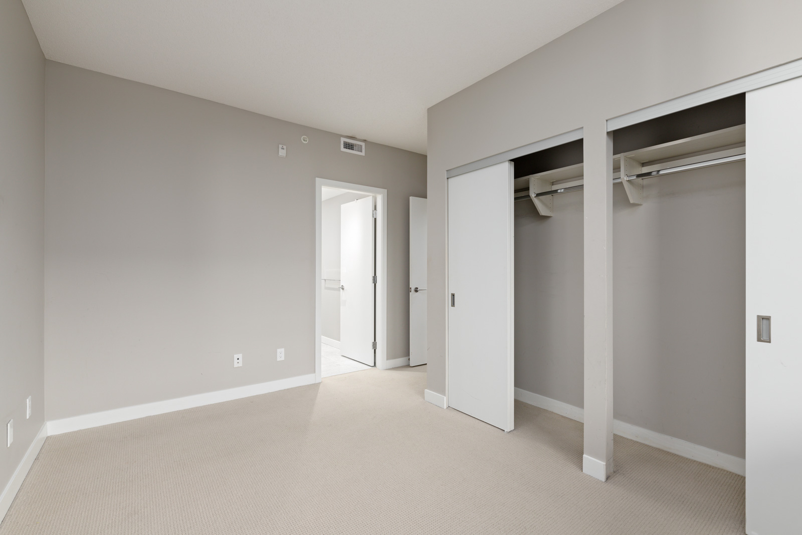 Empty room with beige carpet, gray walls, an open door leading to a bright hallway, and a closet with sliding white doors.