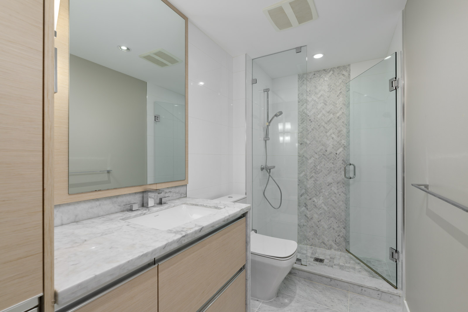 Modern bathroom with a marble countertop, wooden cabinets, wall-mounted sink, glass shower enclosure, and herringbone-patterned shower tiles.