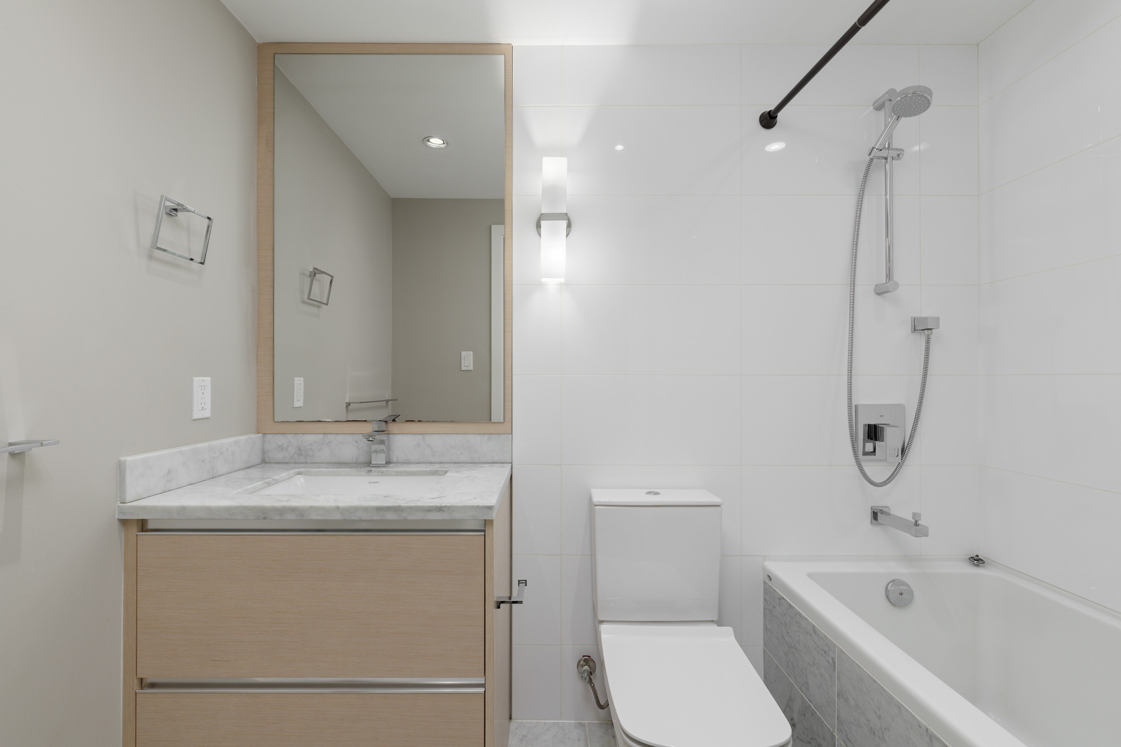 Modern bathroom with light wood vanity, marble countertop, wall mirror, toilet, white tiled walls, bathtub, and shower with overhead fixture.