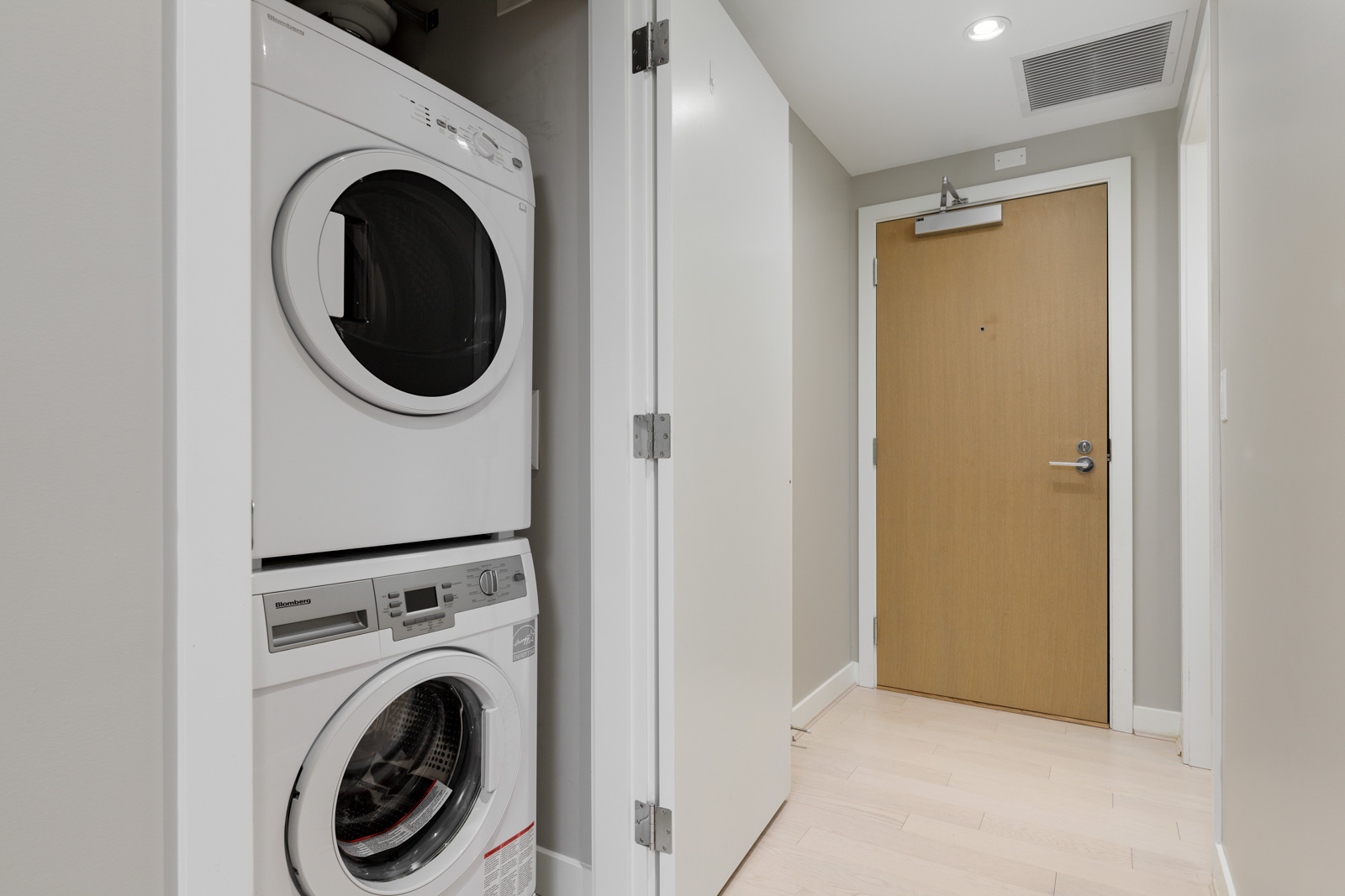 A stacked washer and dryer unit is installed in a small laundry closet next to the front door of a modern apartment.