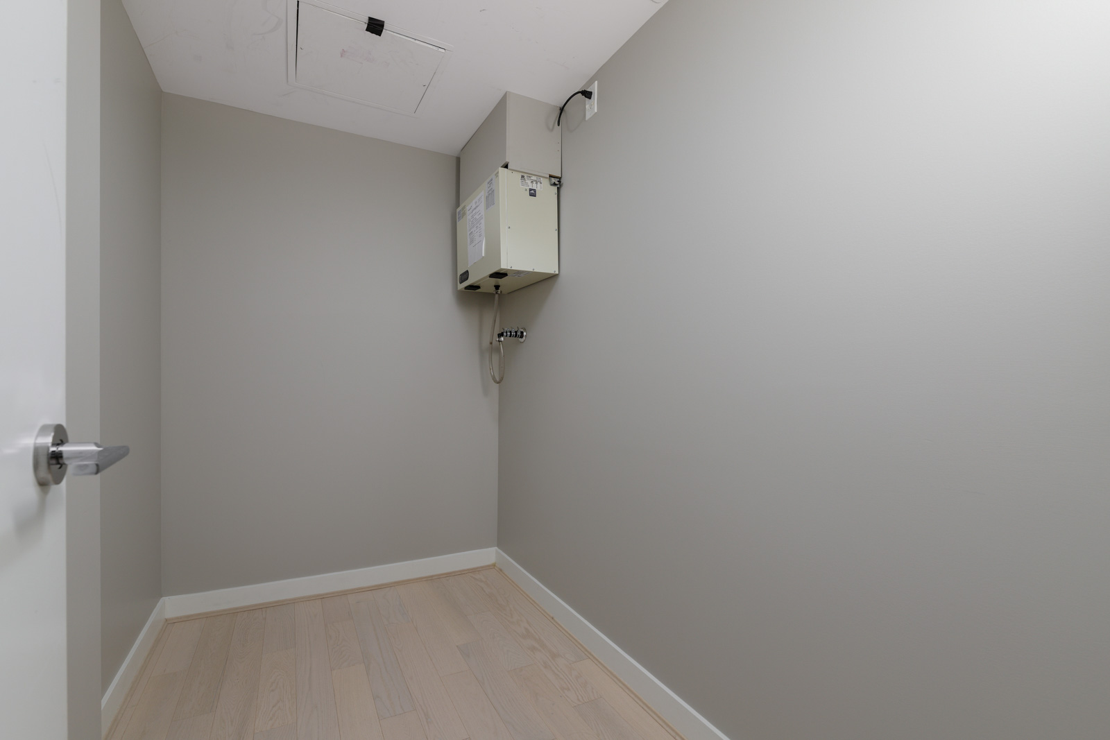 Empty, small utility room with light wood flooring, gray walls, a white access panel in the ceiling, and a wall-mounted utility box with pipes.