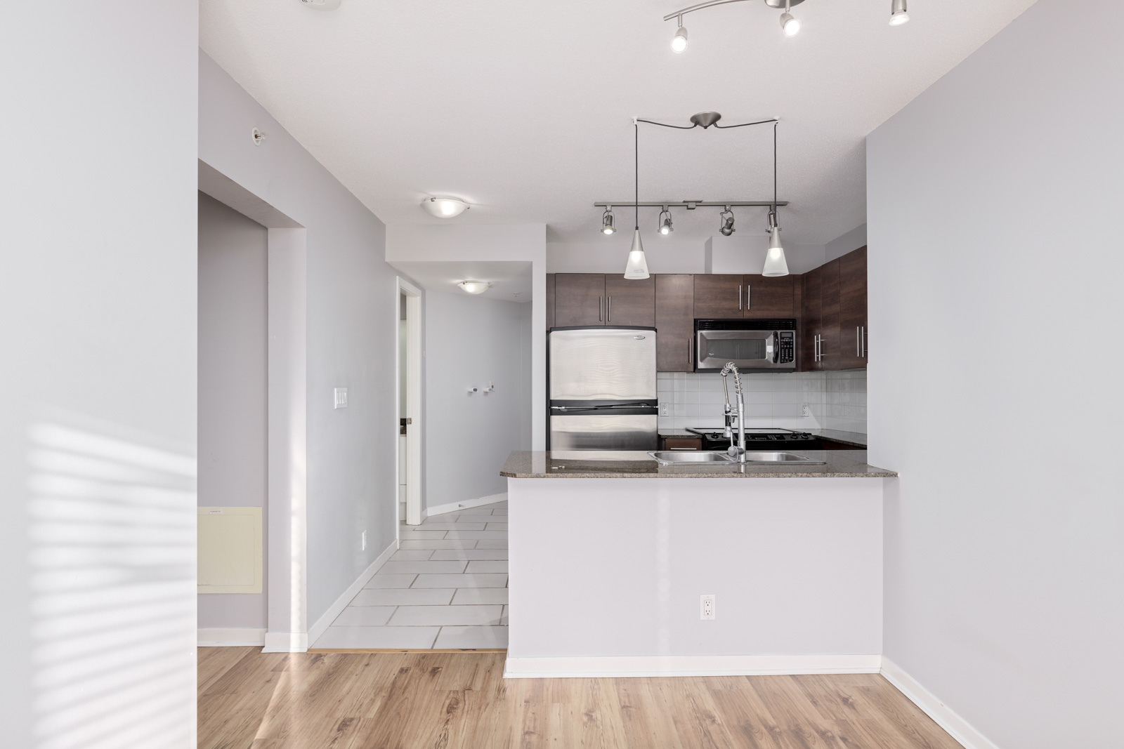 A modern kitchen with stainless steel appliances, dark wood cabinets, a light gray countertop, and hardwood flooring. There is track lighting on the ceiling. The walls are painted light gray.