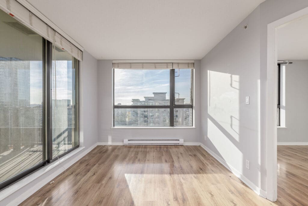 An empty room with large windows, light-colored walls, and hardwood floors. Sunlight streams in from the windows, creating a bright atmosphere.
