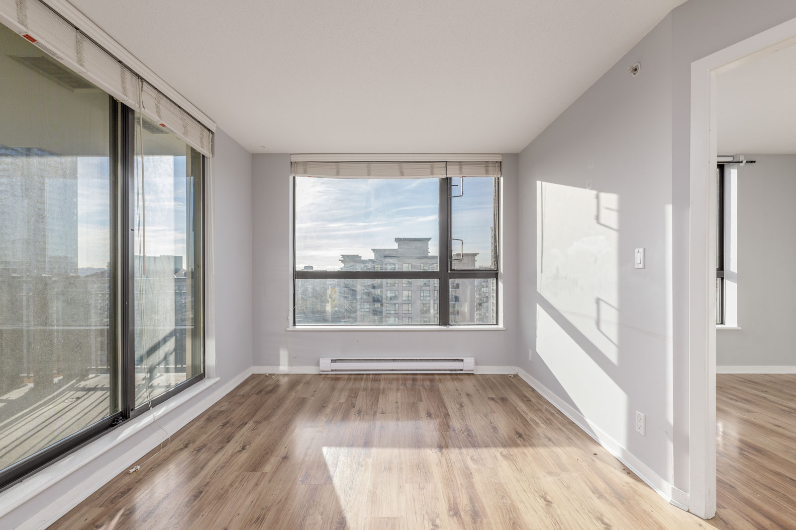 An empty room with large windows, light-colored walls, and hardwood floors. Sunlight streams in from the windows, creating a bright atmosphere.