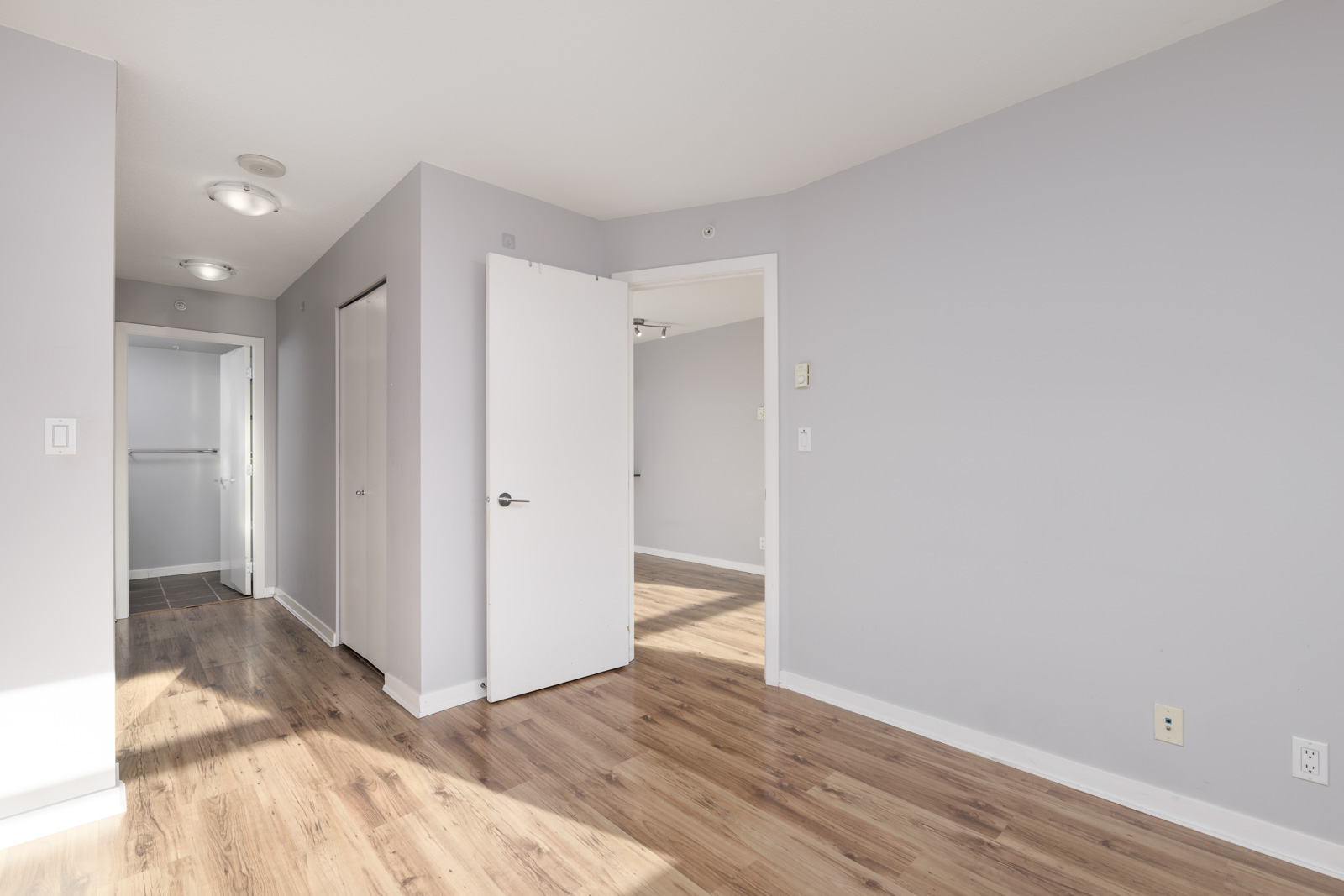 An empty room with light gray walls, wooden floor, and open door leading to another room. Hallway with a closet and a partially open door to a tiled area on the left. Ceiling lights are visible.