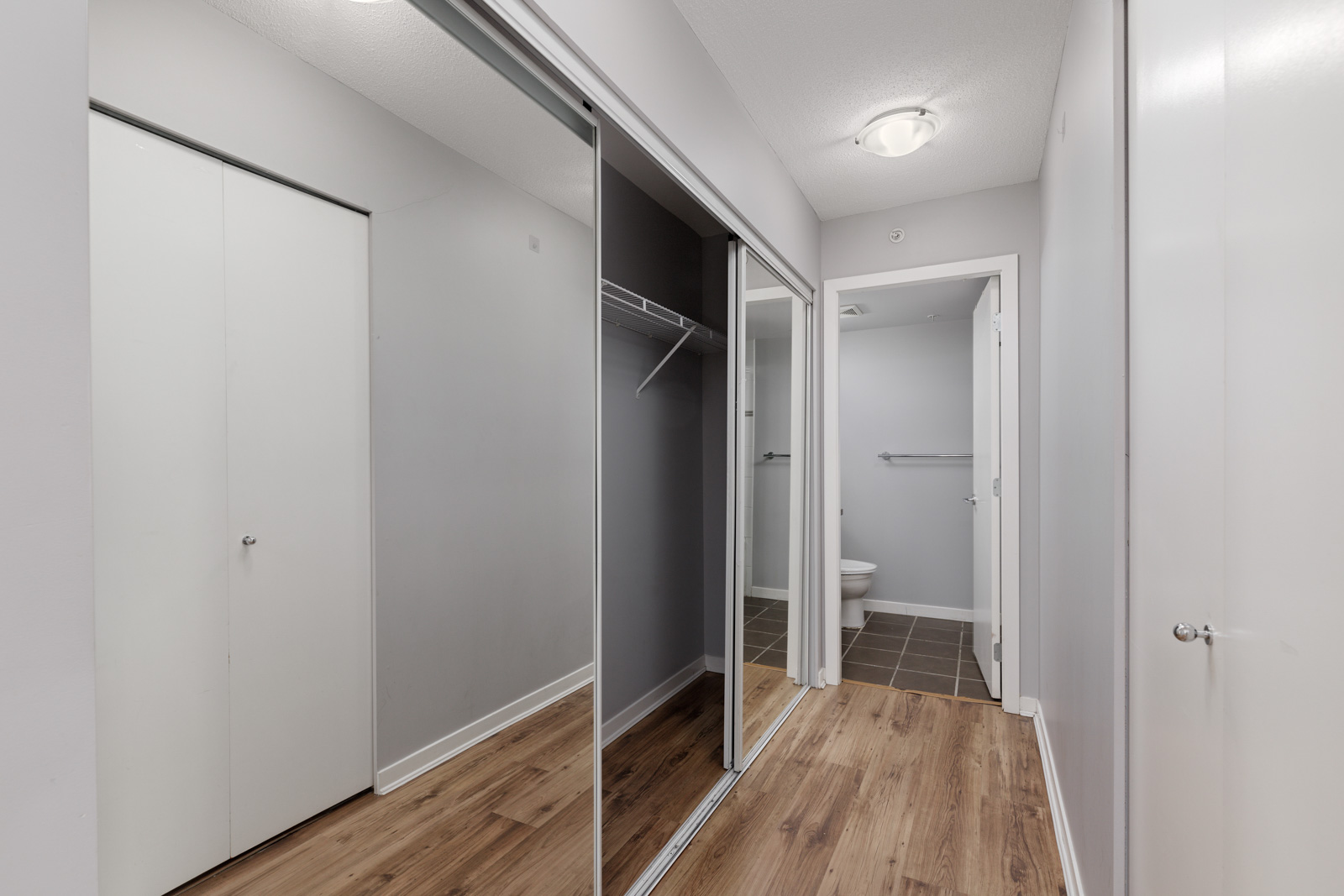 A hallway with wood flooring, mirrored closet doors on the left, and a view into a bathroom at the end. Walls are painted light gray.