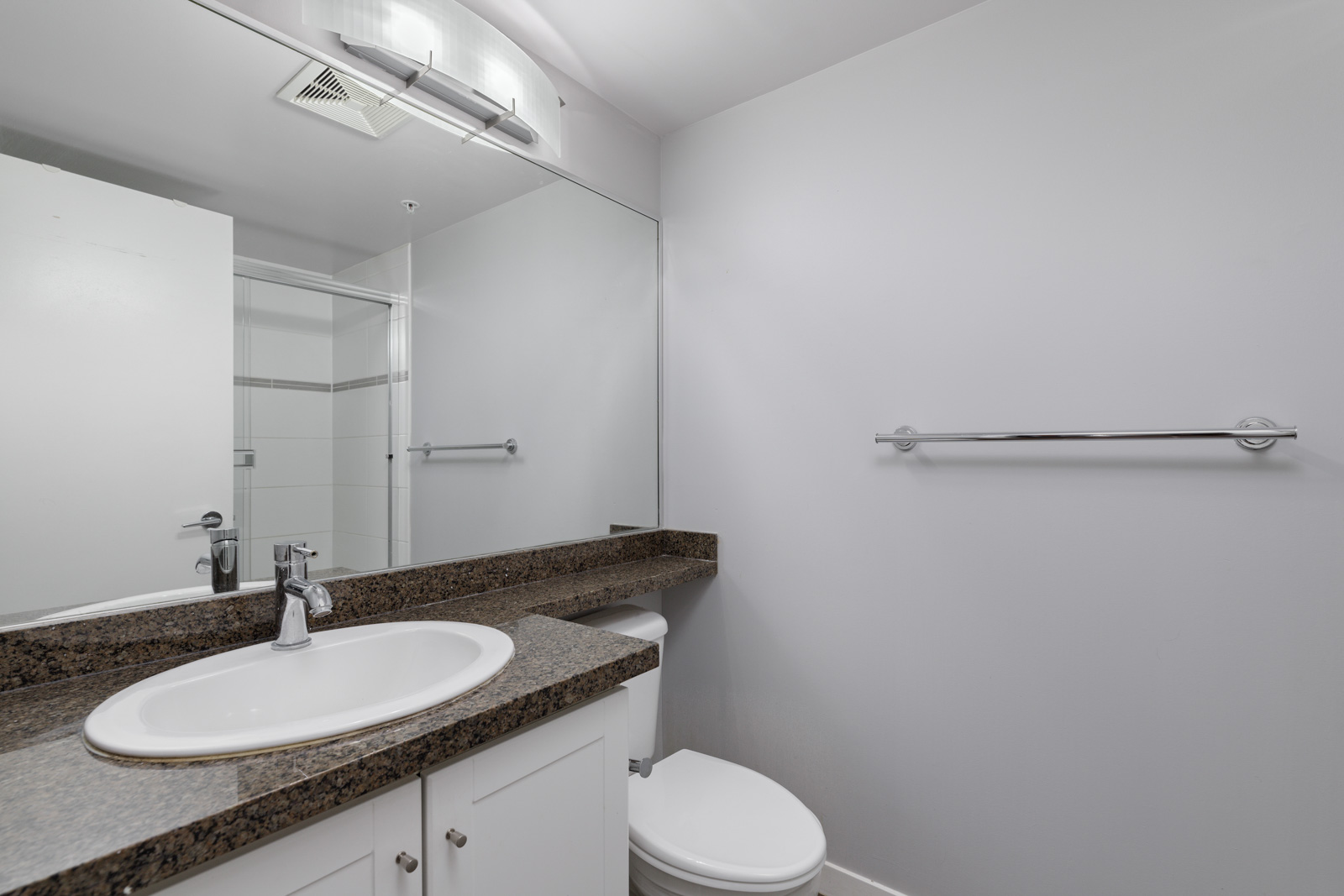 Bathroom with a large mirror, granite countertop, sink, and toilet. Empty towel rack on the right wall. White walls and ceiling.
