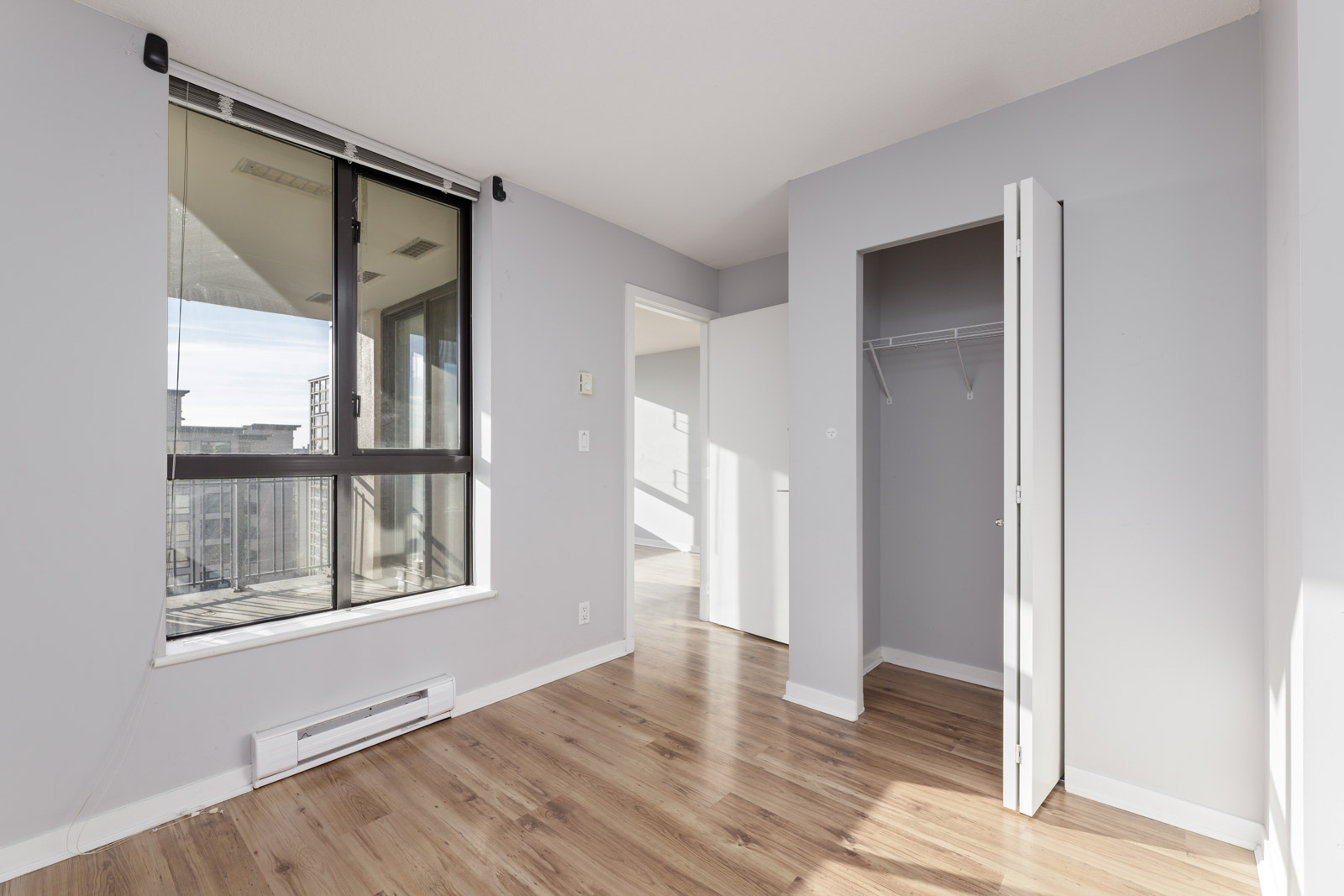 Empty room with light gray walls, a large window, wooden flooring, an open closet, and a white door.