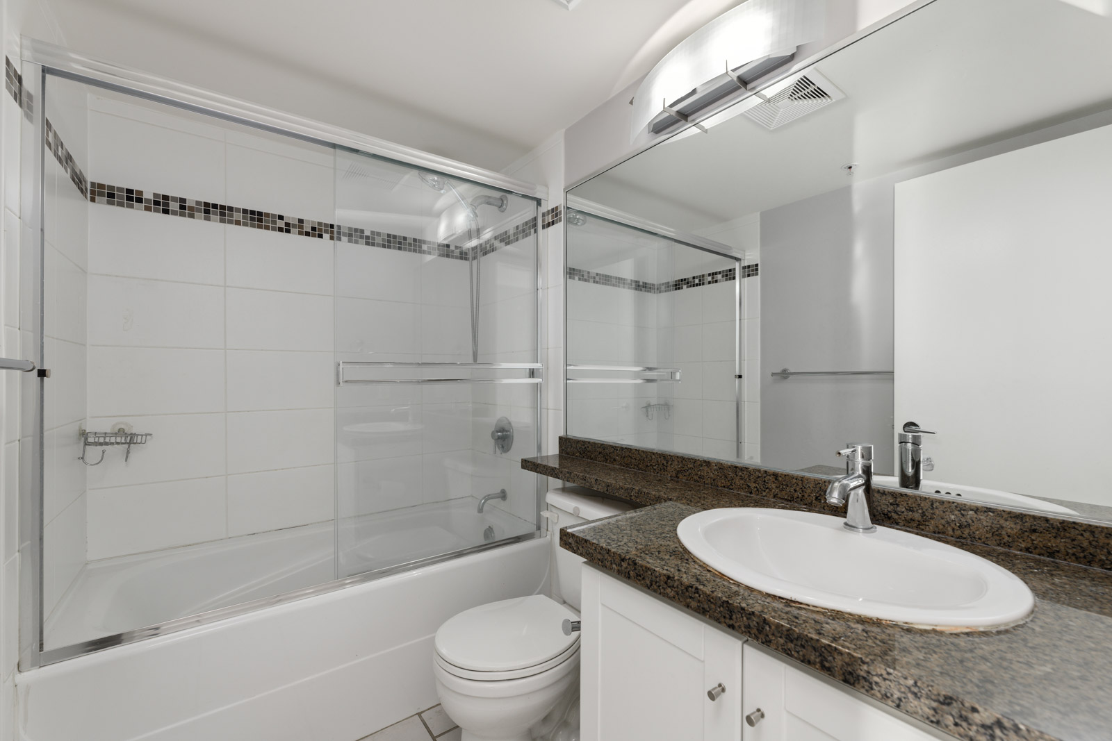 A clean, modern bathroom with a bathtub shower combo, frameless glass door, white tiles, brown granite countertop, sink with silver faucet, toilet, and large mirror.
