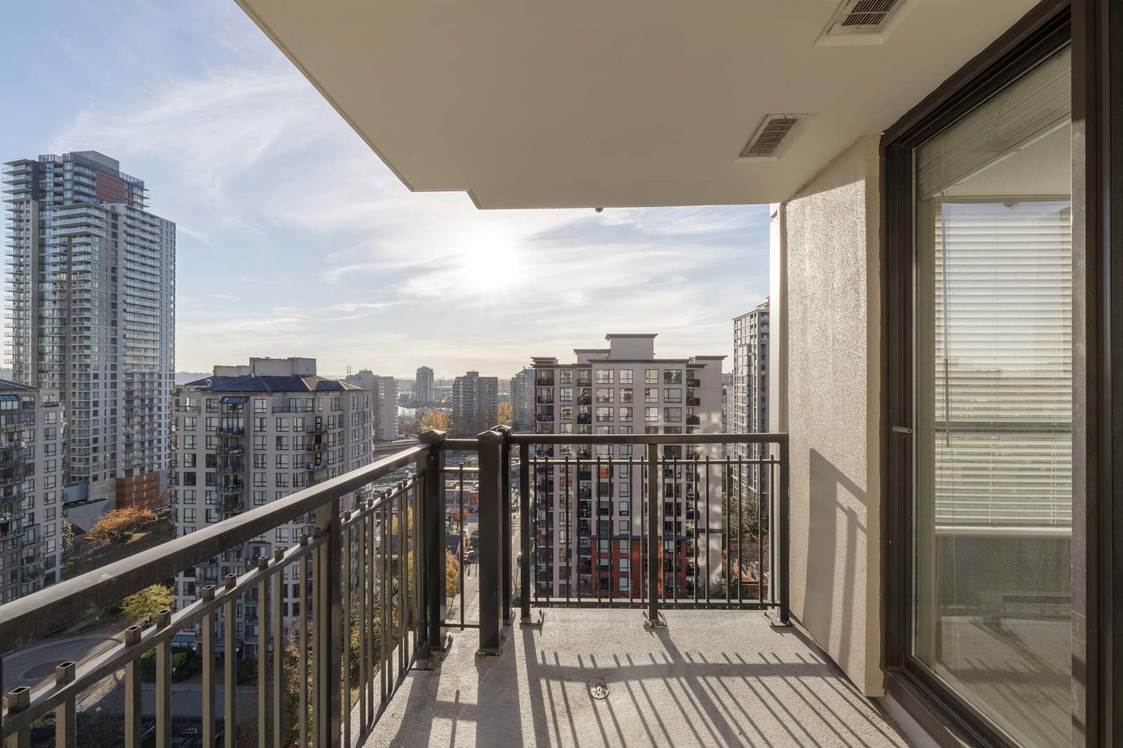 A balcony with metal railings overlooks a cityscape filled with modern high-rise buildings under a sunny sky.
