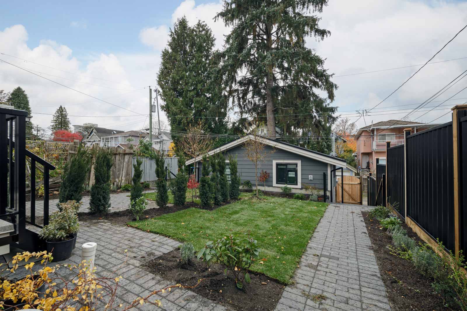 Backyard with paved walkways, a central lawn, small trees, and shrubs; a detached garage and tall evergreen trees are in the background.