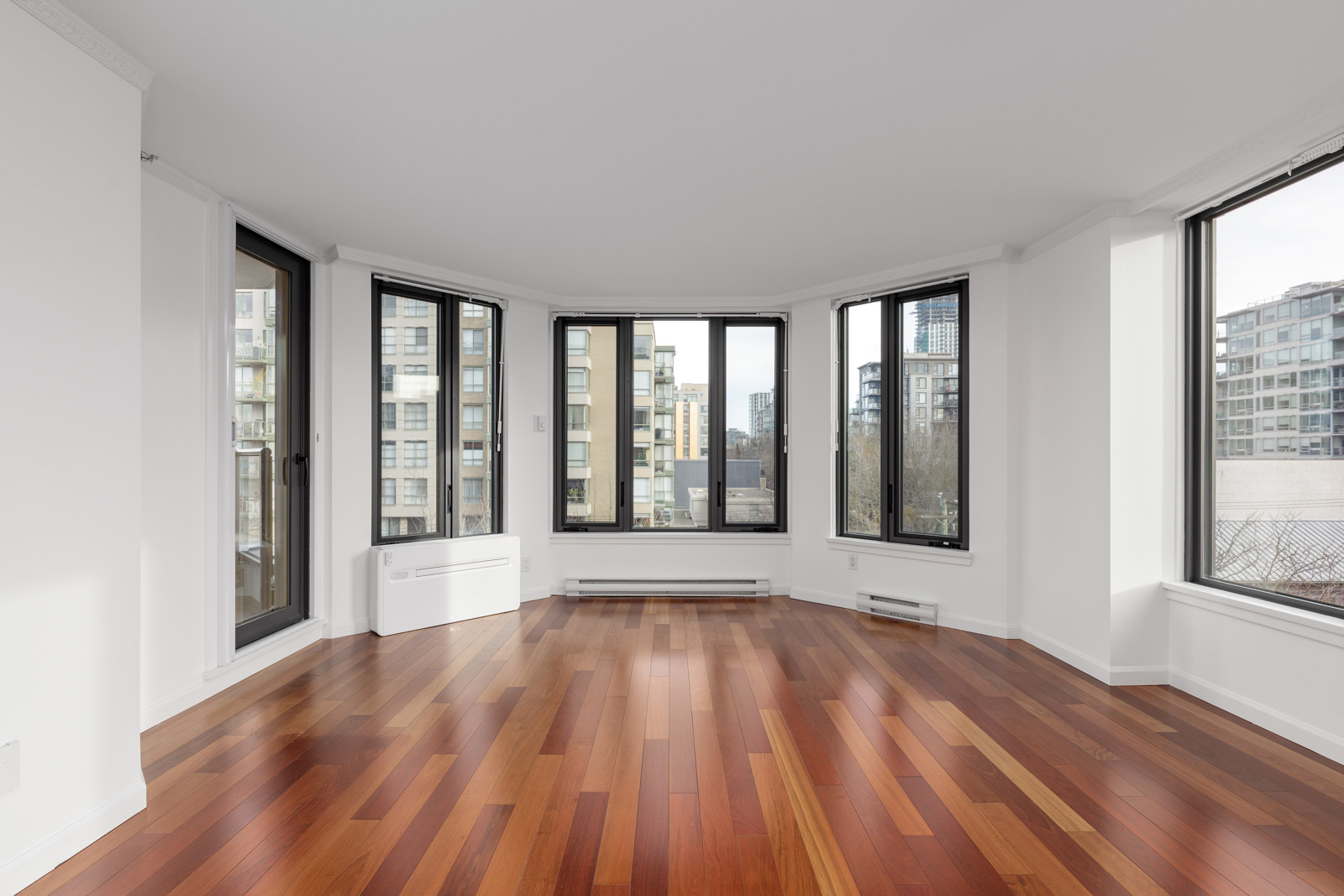 Empty modern apartment room with large windows, white walls, and polished hardwood floors, overlooking city buildings.