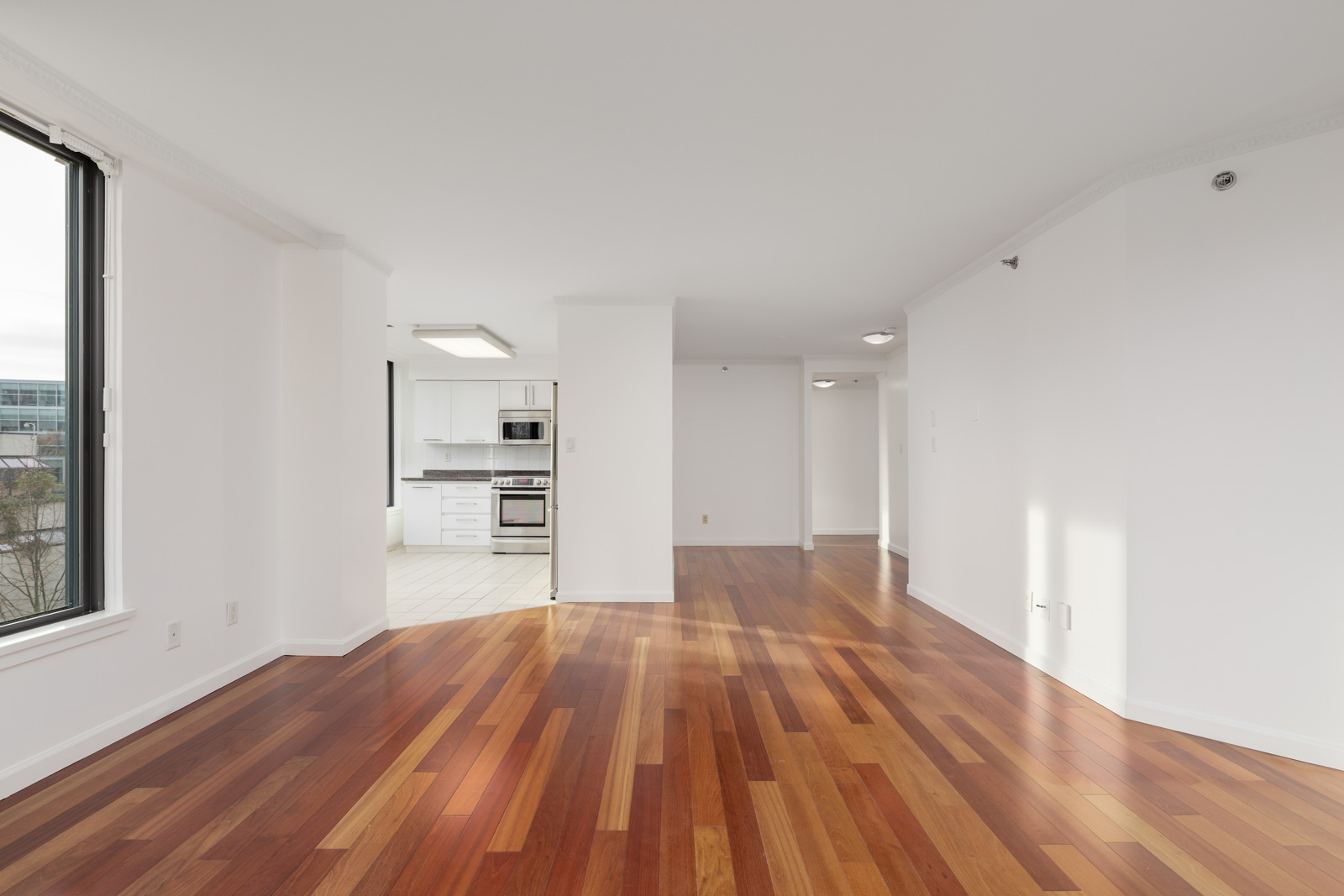 Empty apartment interior with polished wood floors, white walls, large window, and a view into a modern white kitchen with stainless steel appliances.