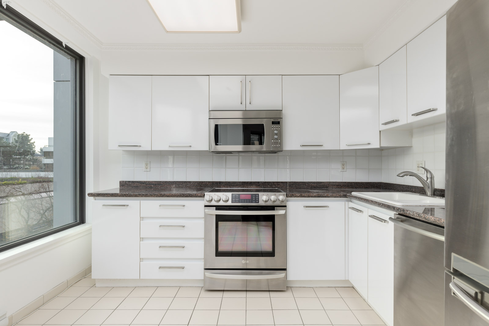 Modern kitchen with white cabinets, stainless steel appliances including an oven and microwave, large window, tiled floor, and granite countertops.