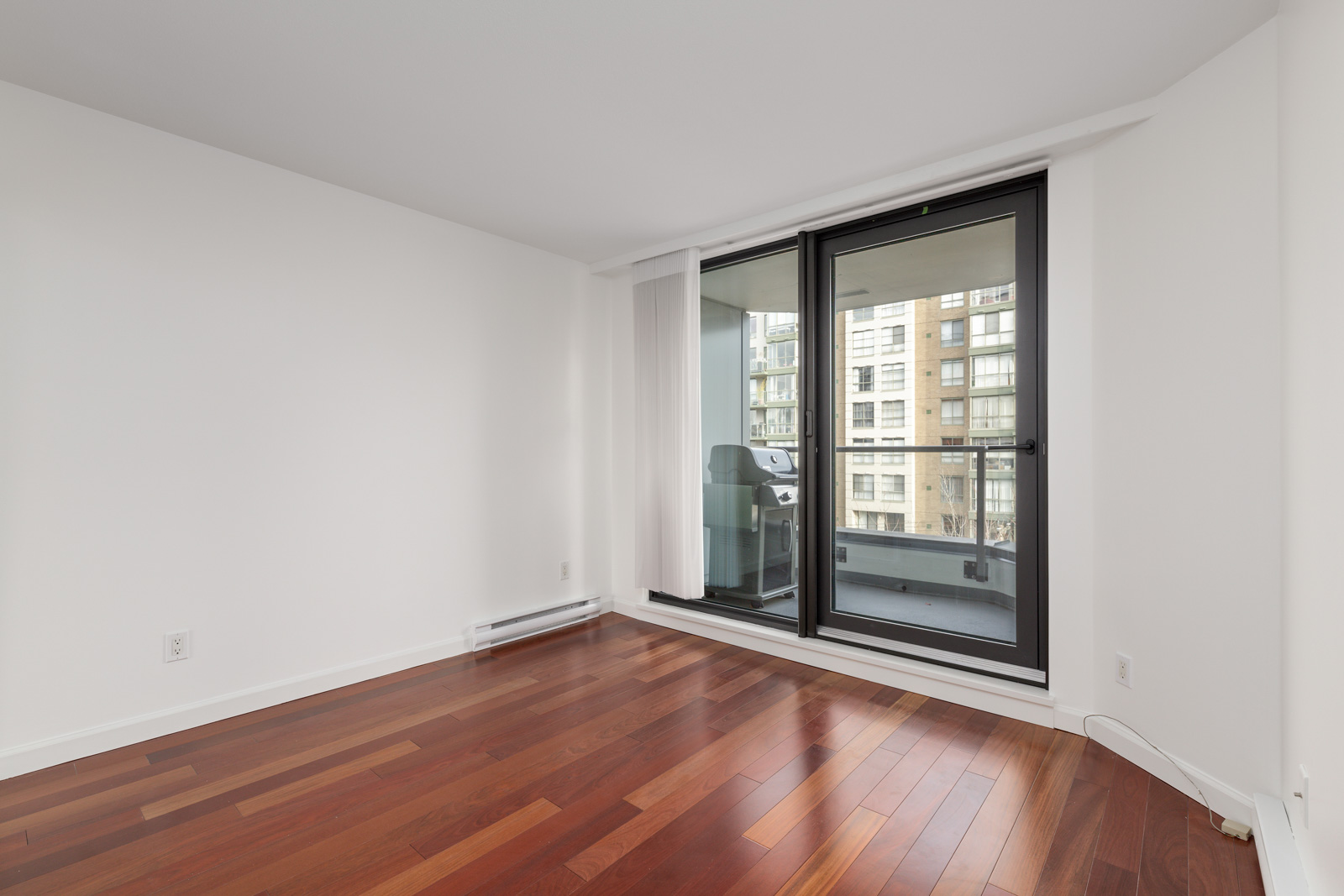 Empty room with white walls, polished wood floor, and large glass doors leading to a balcony with a view of neighboring apartment buildings and a barbecue grill outside.