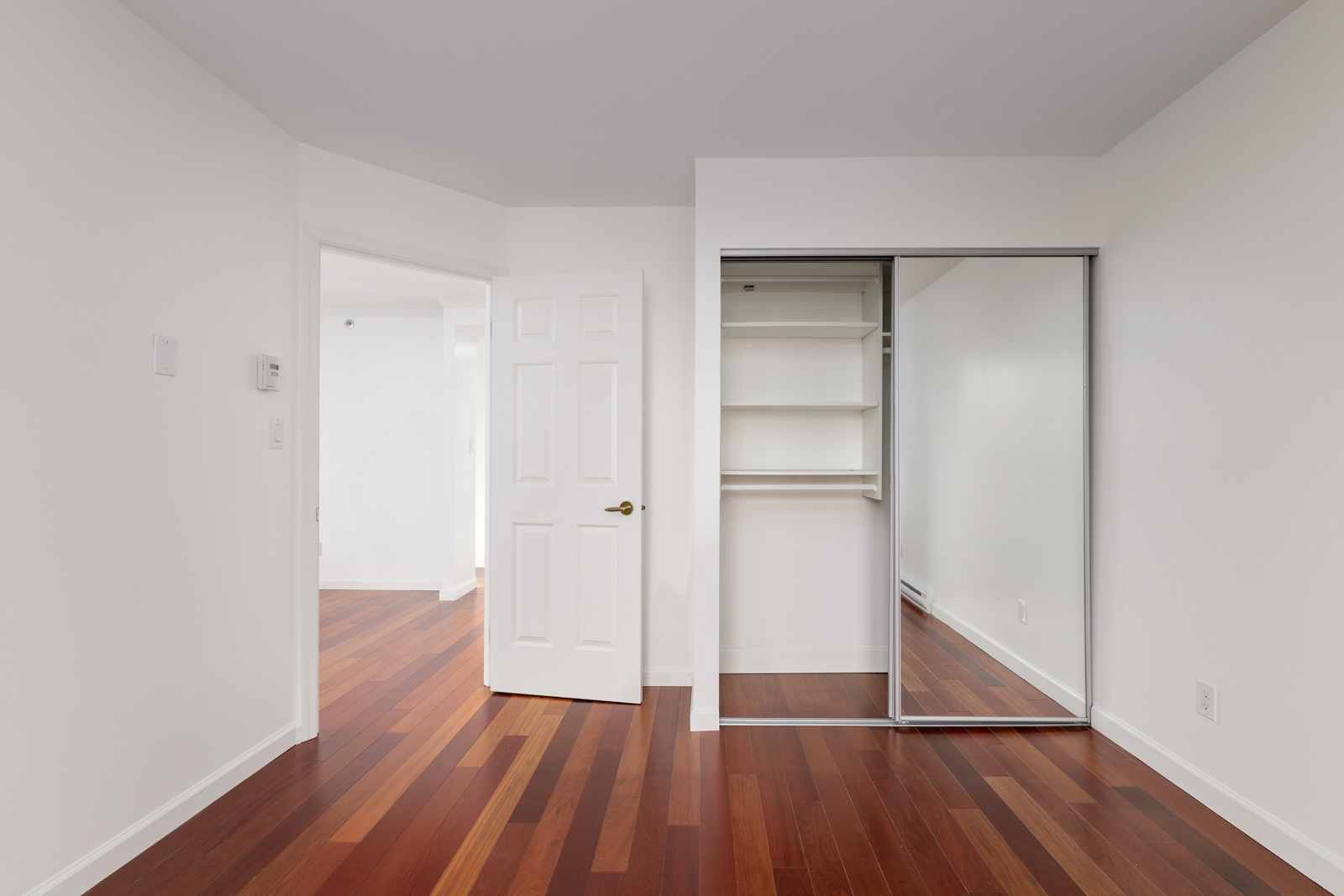Empty room with white walls, an open door, wooden floors, and a built-in closet with sliding mirrored doors and shelves.