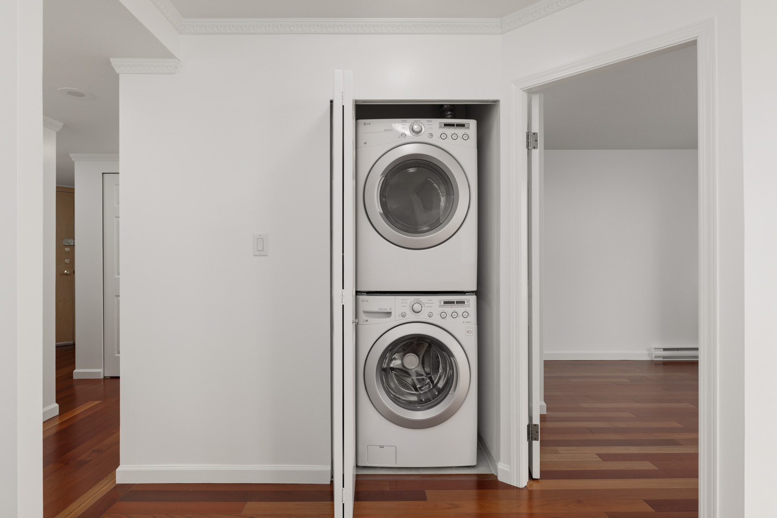 A stacked washer and dryer are installed in a small laundry closet with white doors, in a room with white walls and wood flooring.