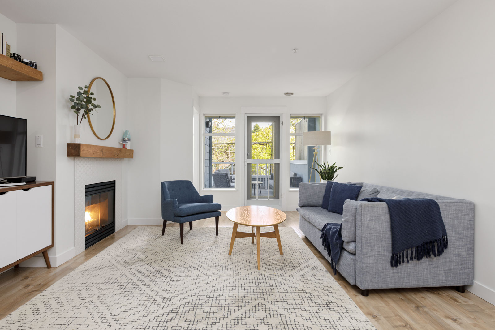Modern living room with a gray sofa, blue armchair, round wooden coffee table, fireplace, wall-mounted TV, and large windows letting in natural light.