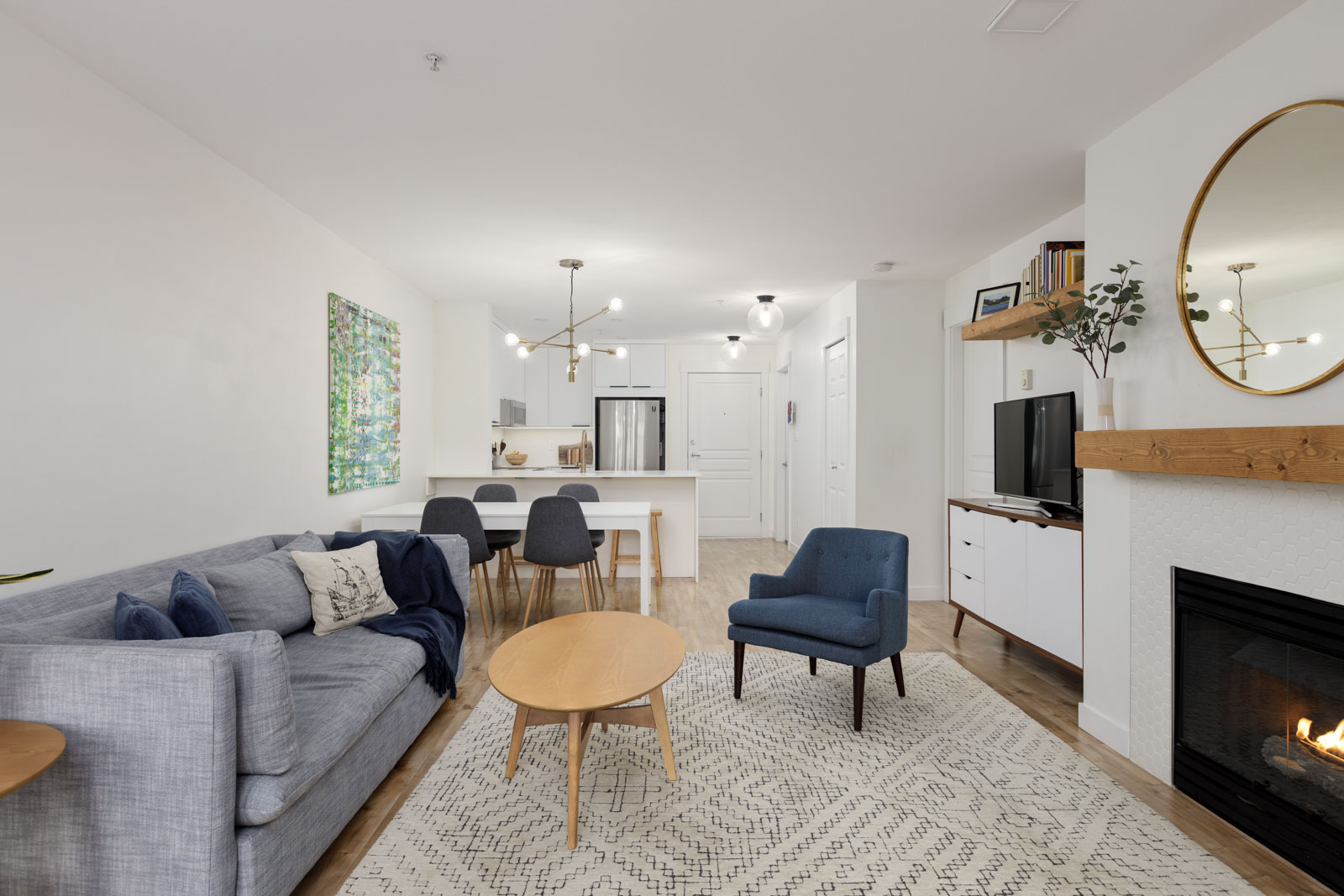 Modern open-concept living room and kitchen with gray sofa, blue armchair, dining table, wall art, fireplace, and mounted TV in a bright, neutral color scheme.