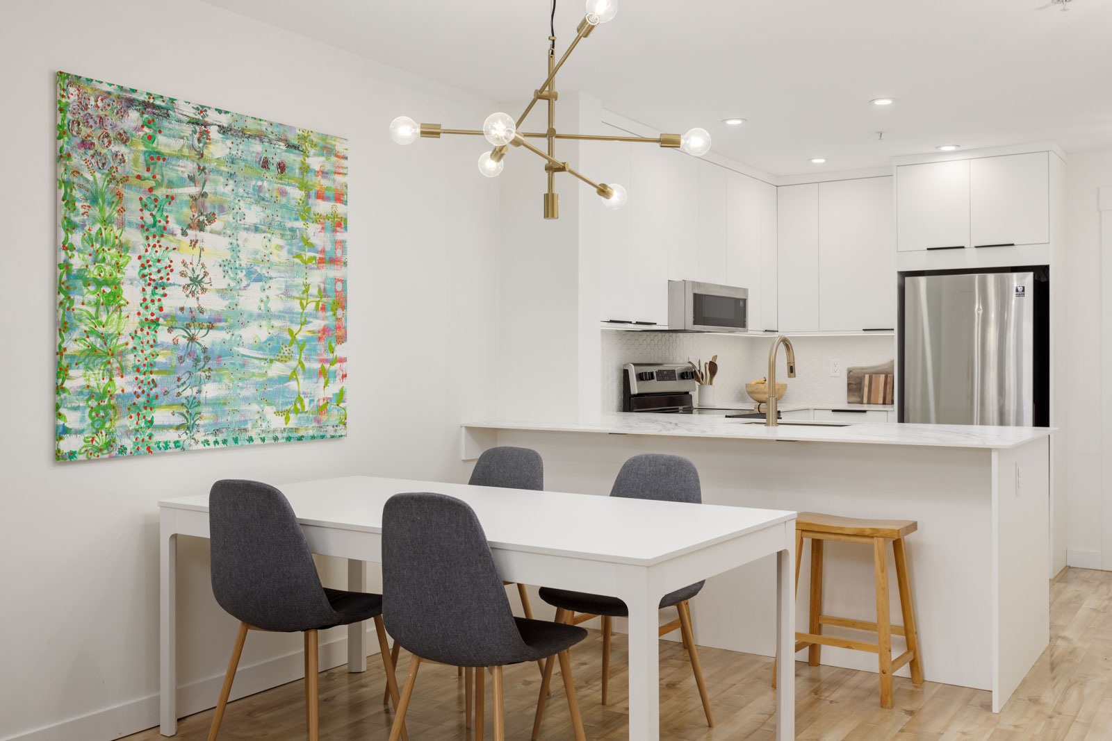 A modern kitchen and dining area with white cabinets, a white dining table, four dark chairs, a wooden stool, abstract wall art, and a brass light fixture.