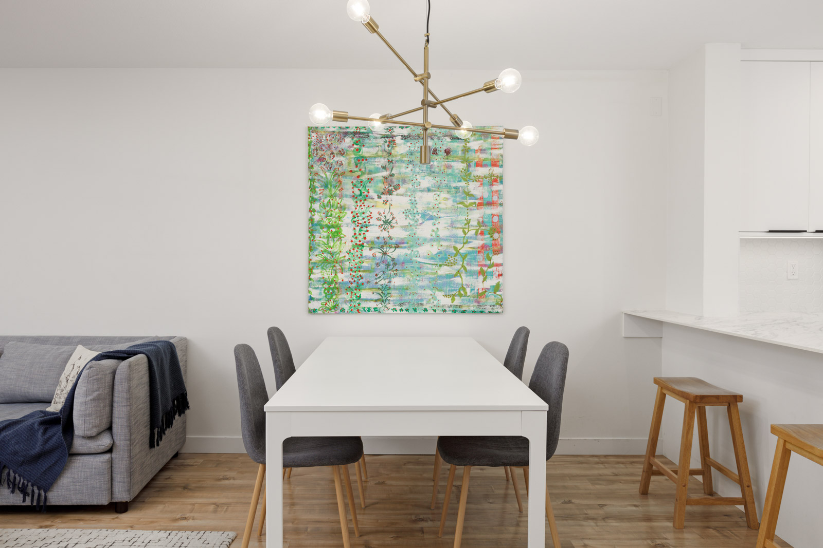 A modern dining area with a white table, four gray chairs, abstract artwork on the wall, a gold chandelier, and a sofa and stools in the background.