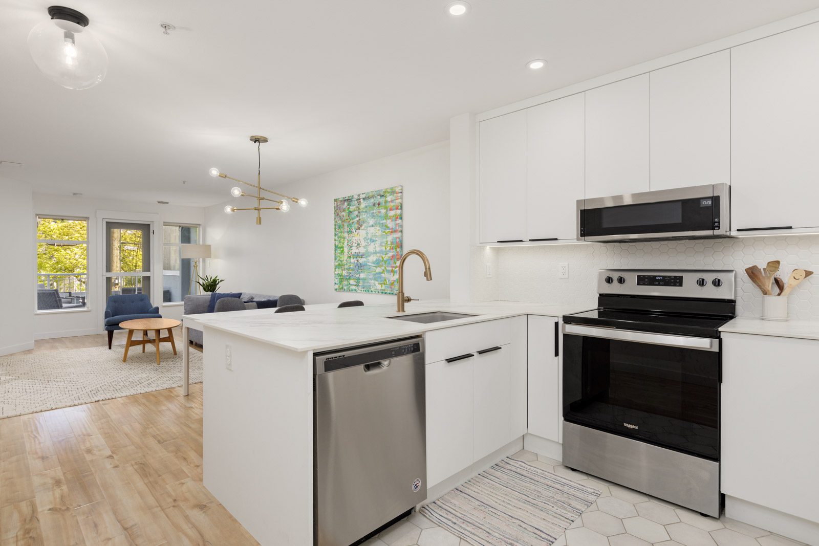 Modern kitchen with white cabinets, stainless steel appliances, and a peninsula counter, open to a bright living area with wood flooring and large windows.