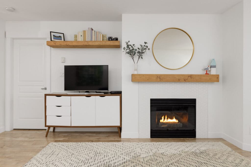 Modern living room with a white cabinet and TV on the left, a fireplace with a round mirror above on the right, and wood shelves decorated with books and small objects.