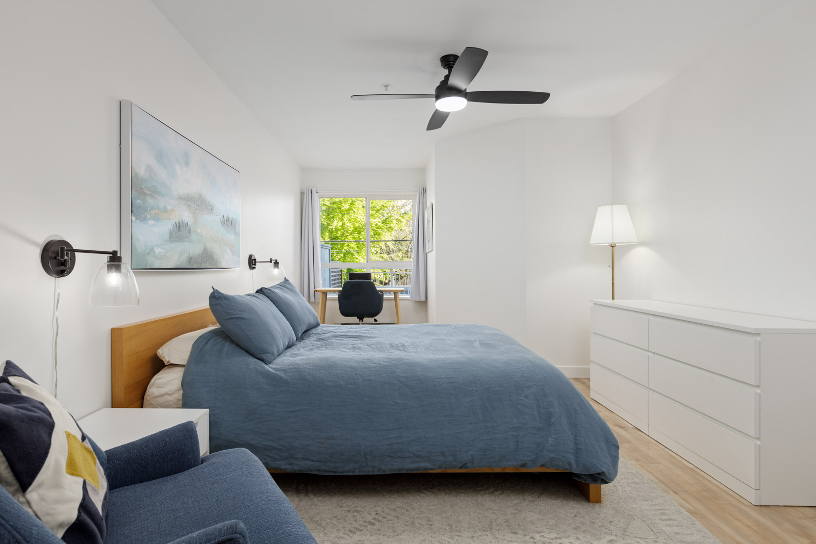 A modern bedroom with a blue bed, white dresser, armchair, desk, and window overlooking trees, featuring a ceiling fan and minimalist decor.