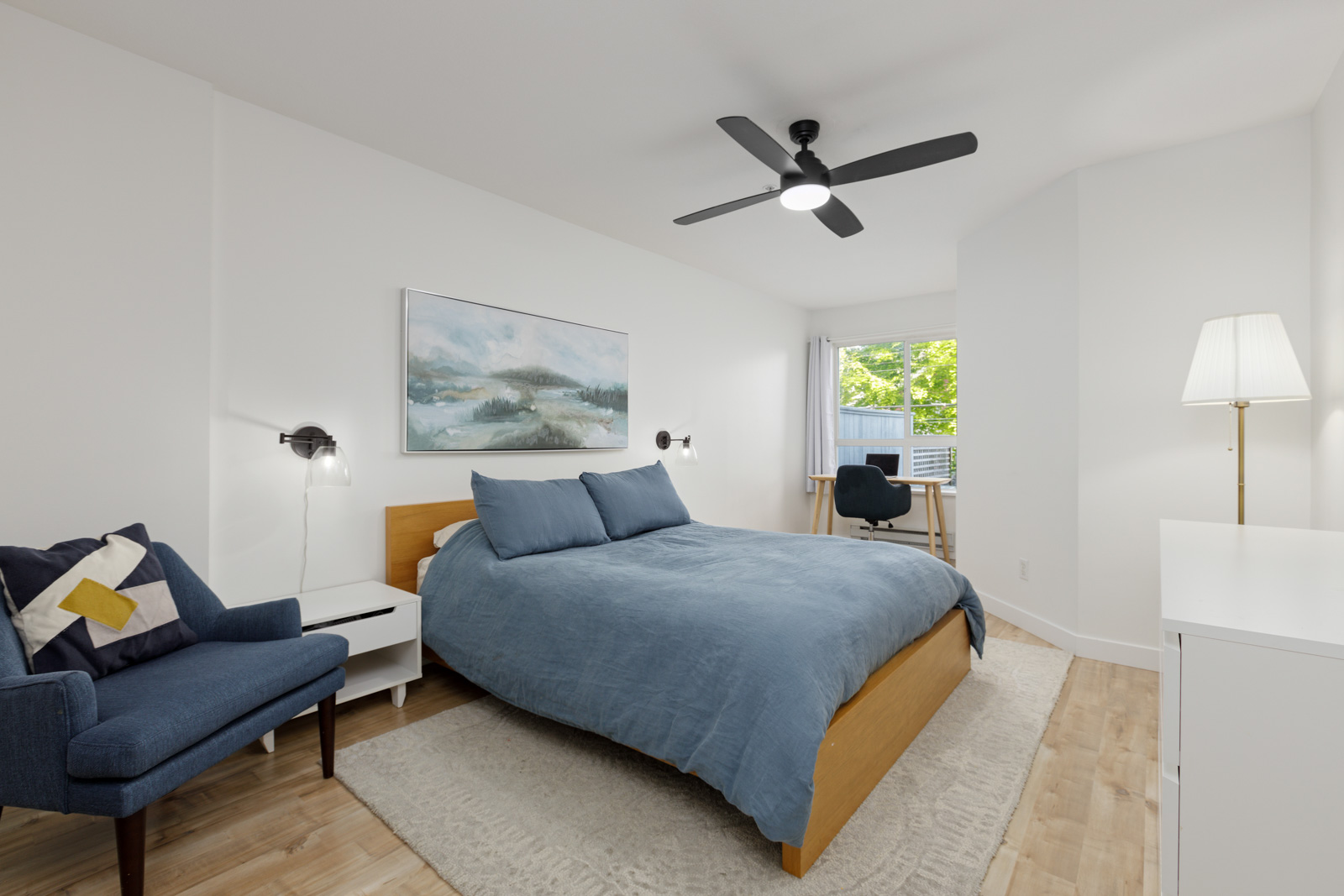 A modern bedroom with a wooden bed, blue bedding, armchair, desk, ceiling fan, and a large window with natural light.