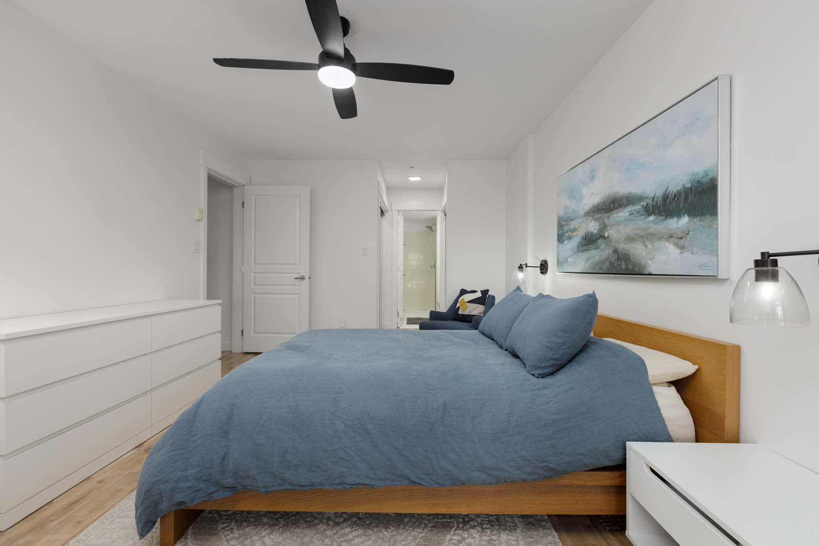 Modern bedroom with a wooden bed frame, blue bedding, white walls, a ceiling fan, dresser, and wall art above the bed. A chair and a doorway are visible in the background.