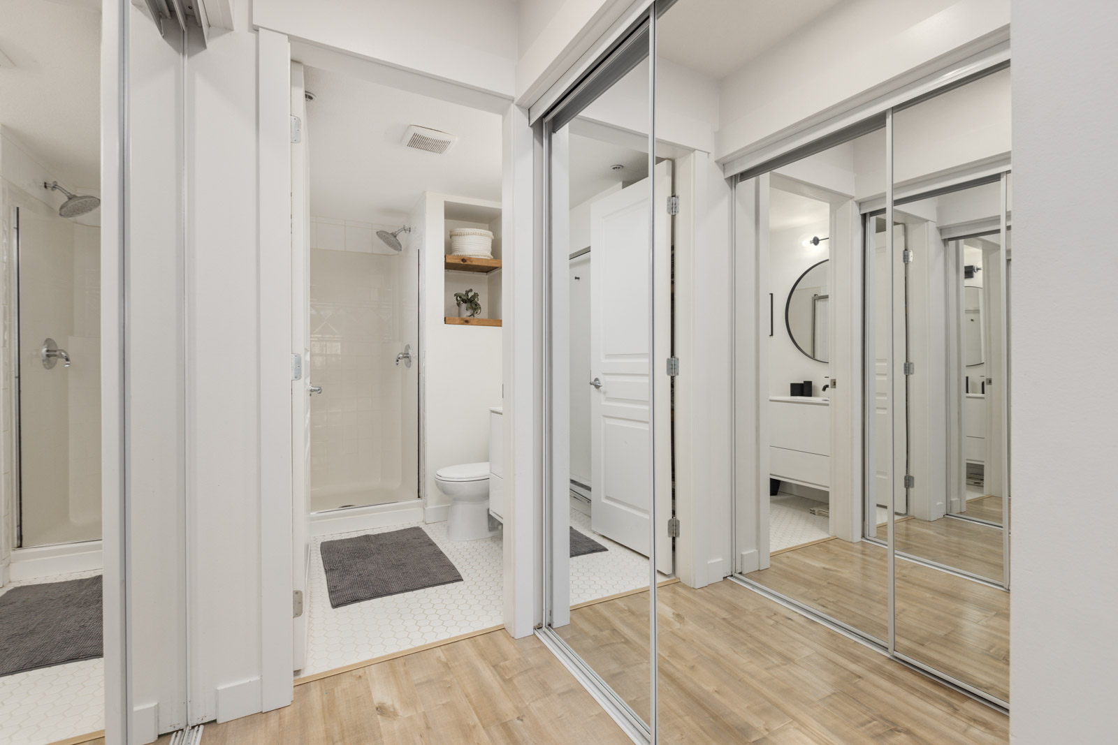 Modern bathroom with a shower, toilet, vanity, and mirrored closet doors reflecting the room; neutral color scheme with wood and tile flooring.