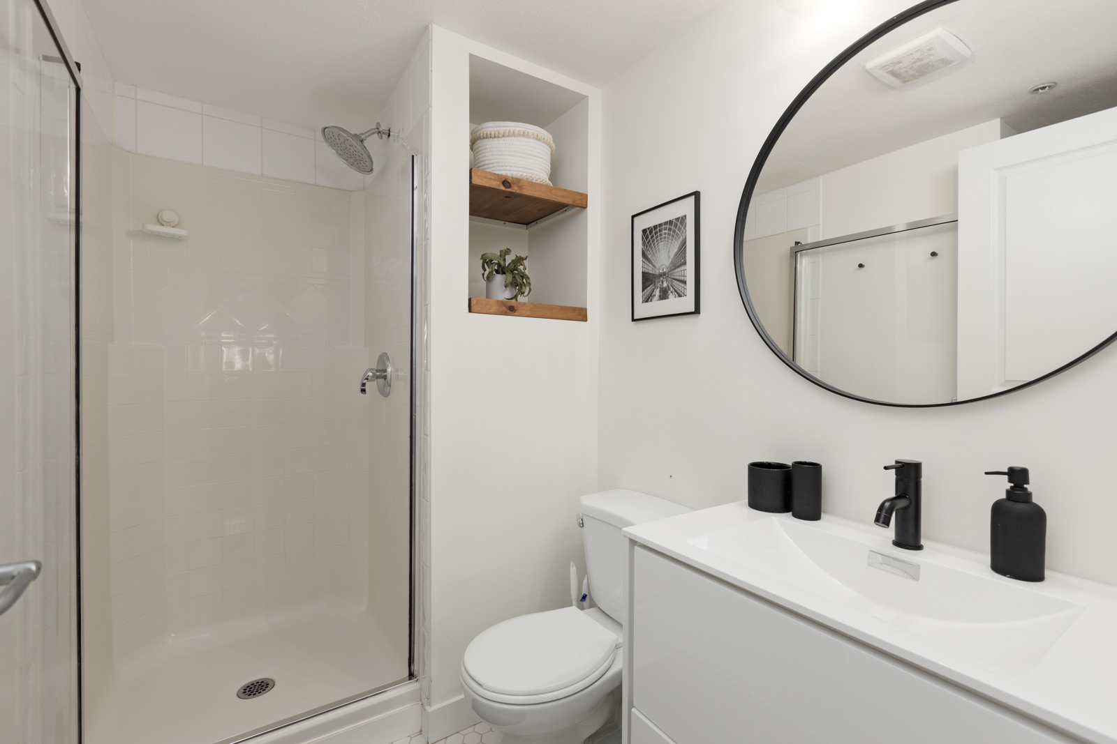 A modern bathroom with a glass shower, white toilet, white vanity with black fixtures, wall shelves, a round mirror, and a framed photo.