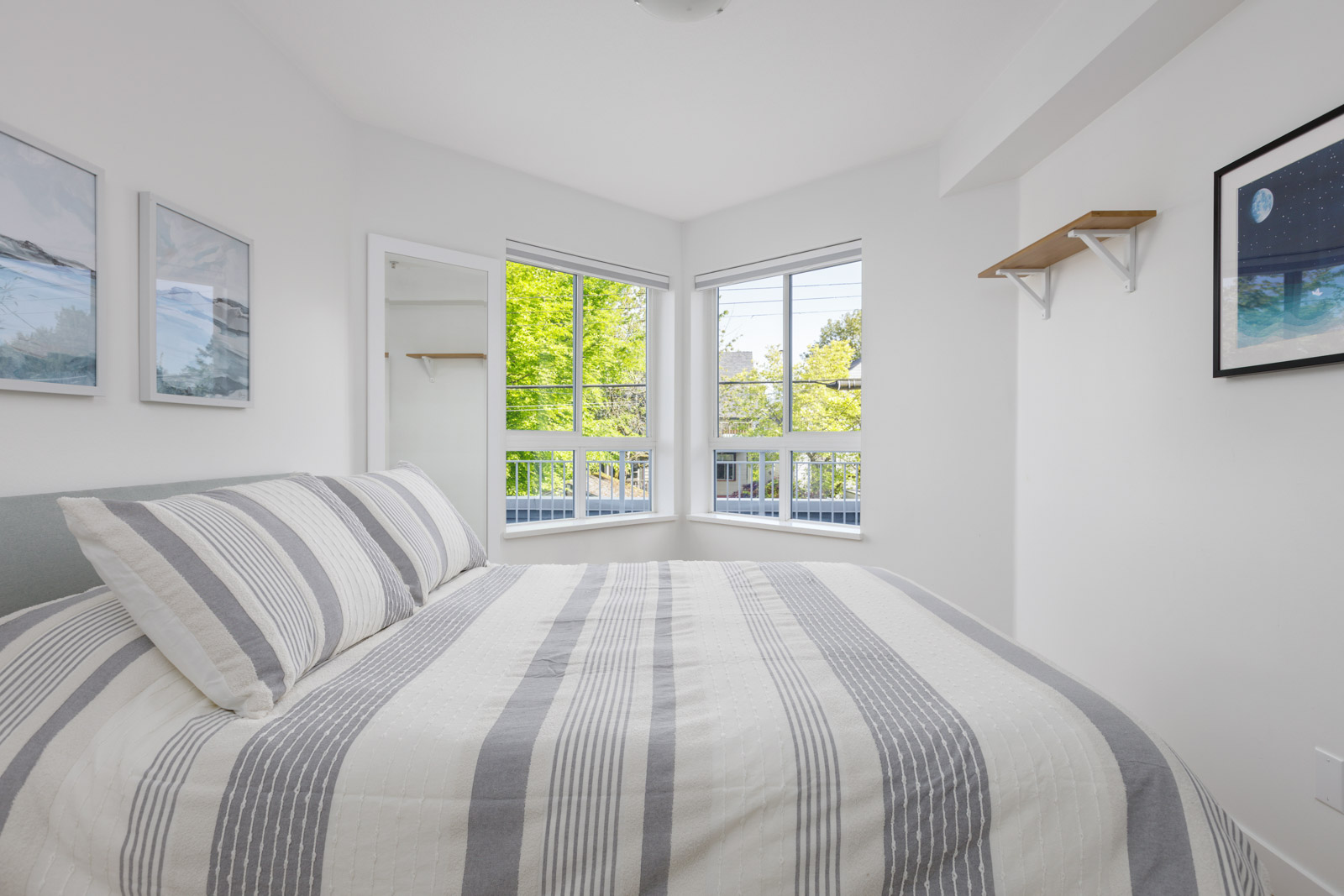 Minimalist bedroom with a striped bedspread, corner windows letting in natural light, white walls, and framed art on the walls.