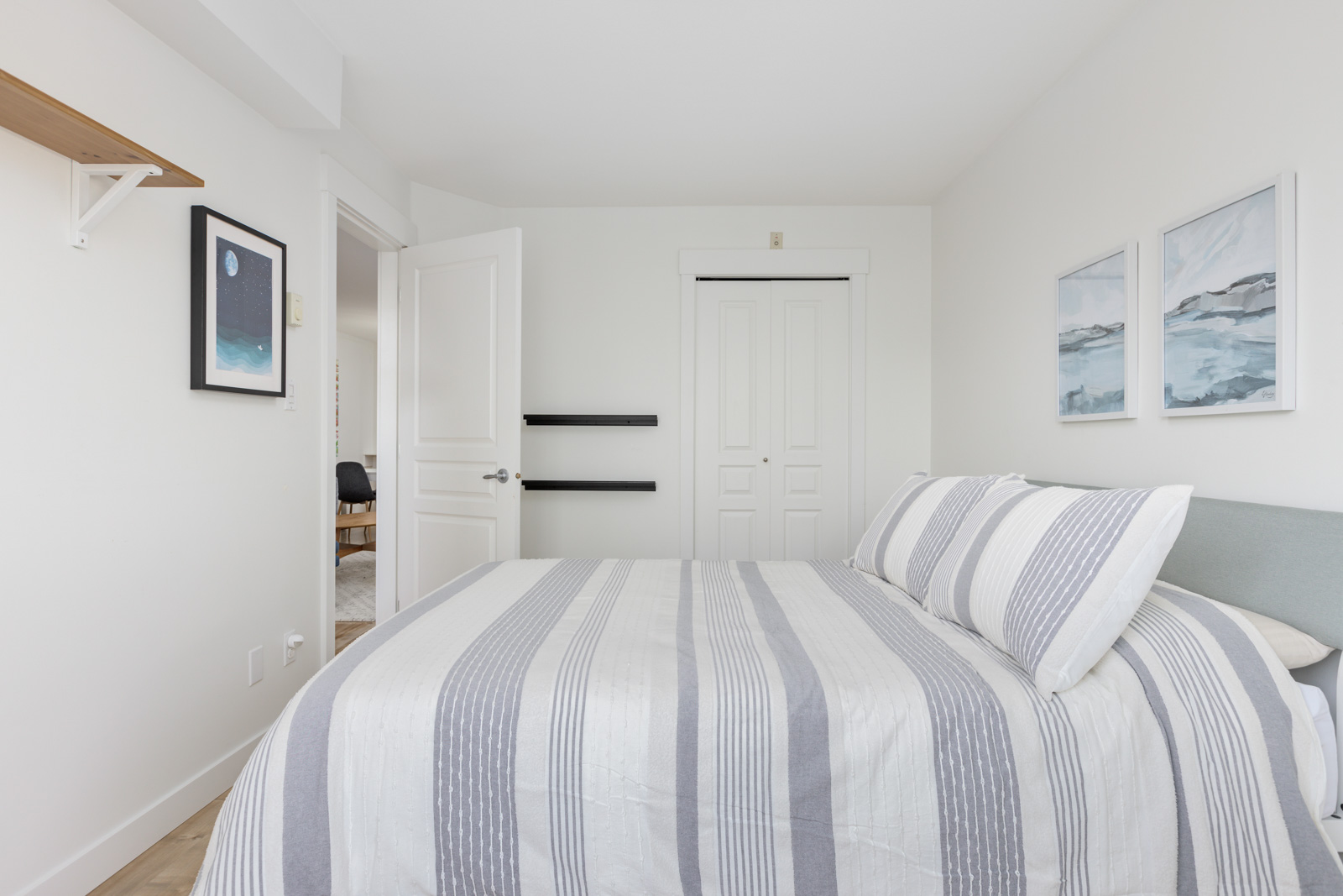 A neatly made bed with striped bedding in a white bedroom featuring minimal decor, a closet with double doors, and framed art on the walls.
