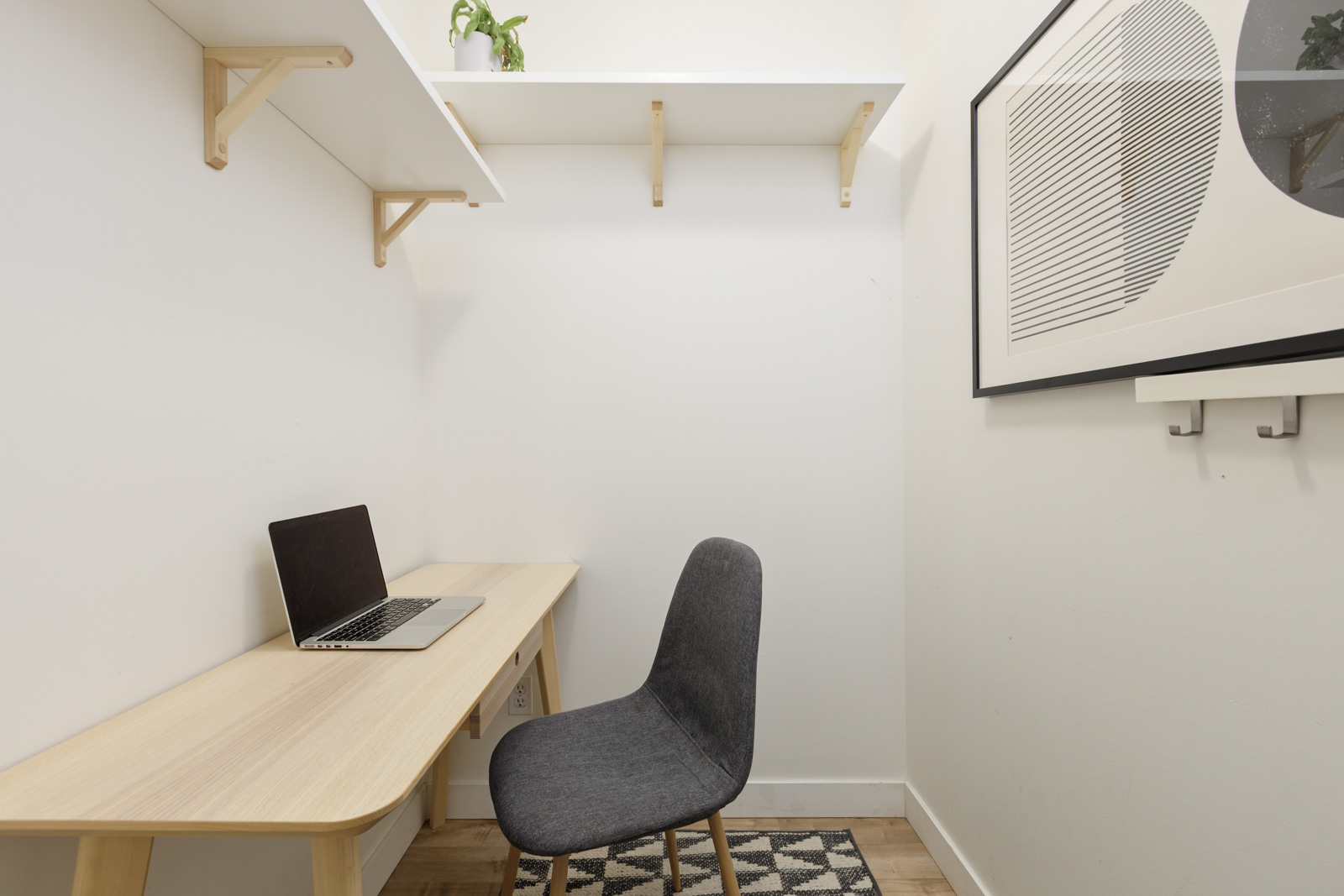 A small home office with a wooden desk, laptop, gray chair, geometric rug, wall shelves, and abstract framed art.