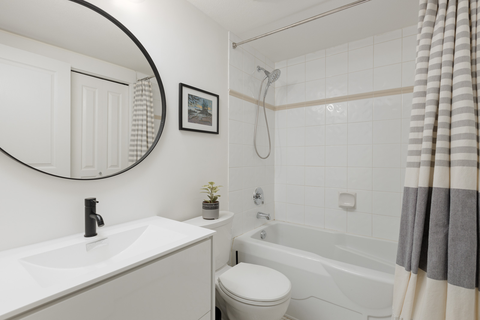 Modern bathroom with a white vanity, round mirror, bathtub with shower, striped shower curtain, framed wall art, and small potted plant.