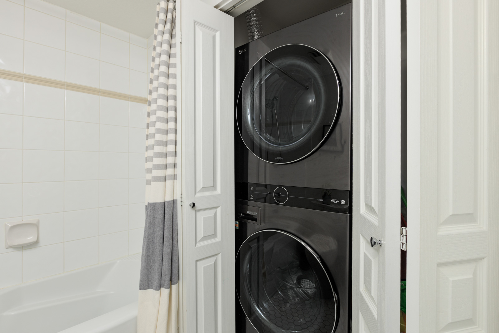 A black stacked washer and dryer set is placed in a closet next to a bathtub with a striped shower curtain in a bathroom.